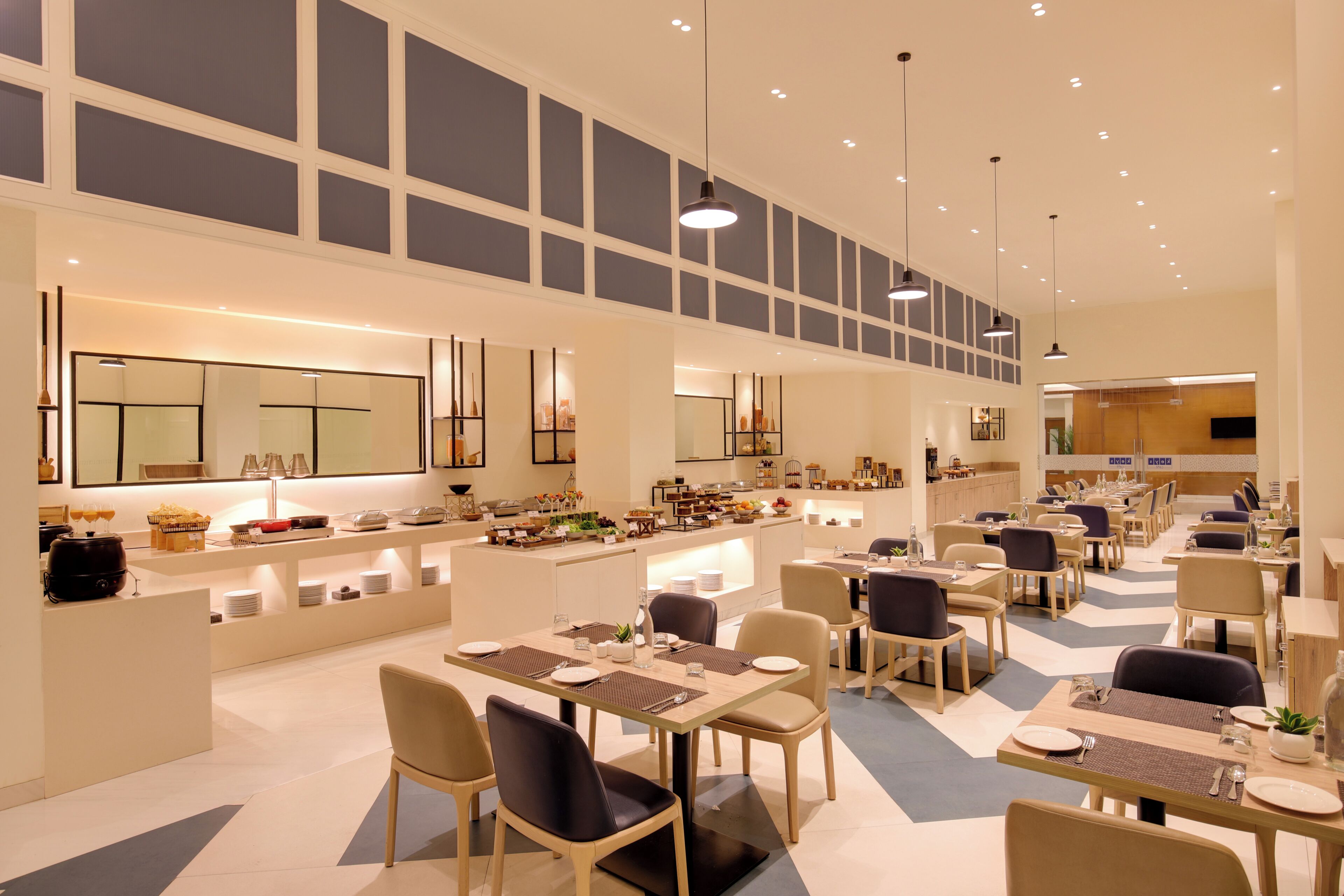 Un amplio comedor de hotel con numerosas mesas y sillas, una barra central con una variedad de alimentos, y grandes ventanales que permiten la entrada de luz natural.