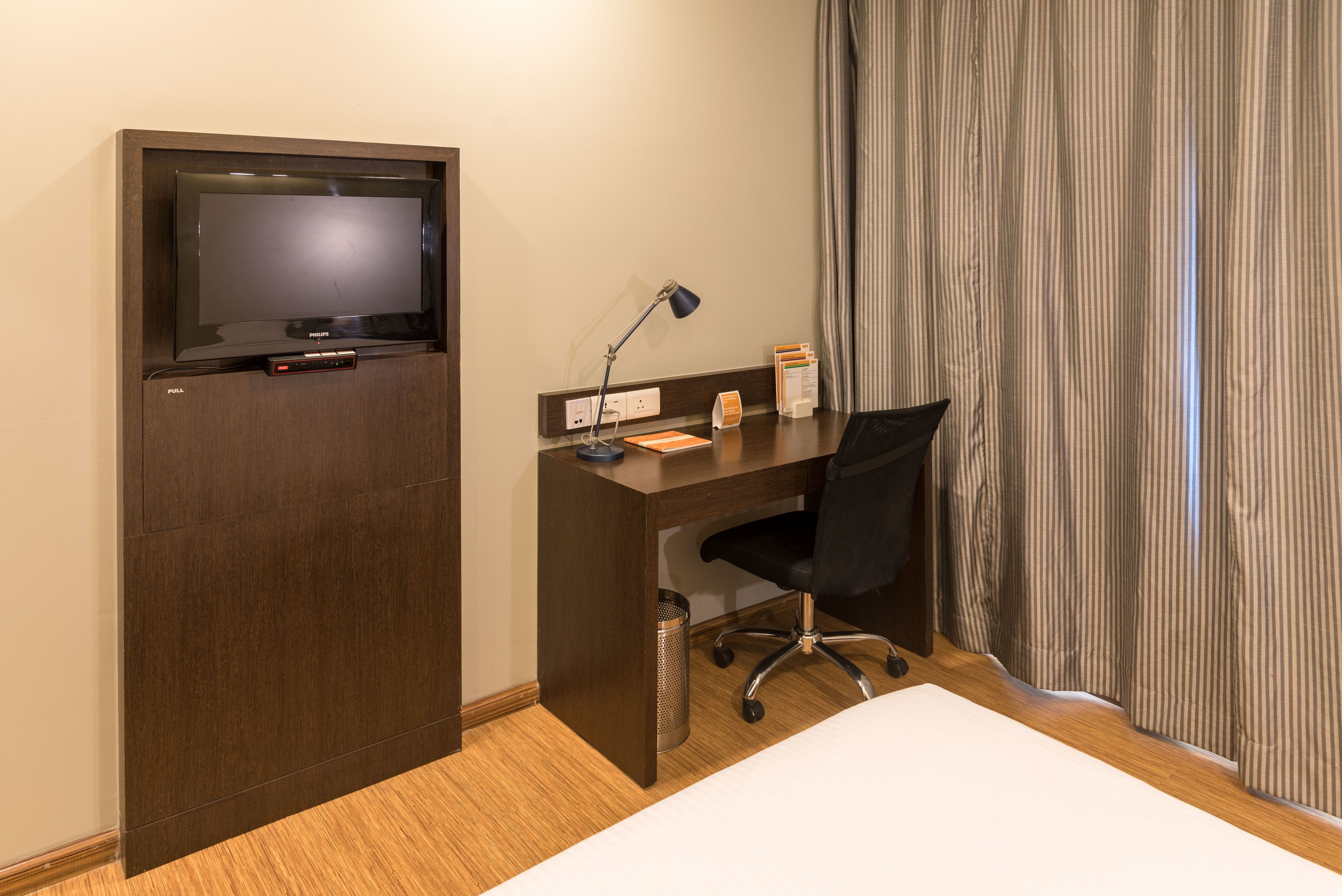 Una habitación de hotel con un escritorio de madera, una silla de oficina negra, un televisor, una lámpara y una cama.