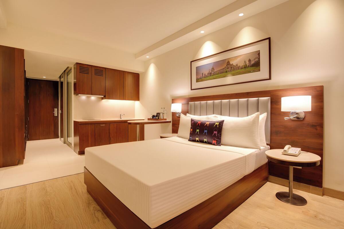 Quarto de hotel com uma cama grande, abajures, uma mesinha com telefone, um quadro na parede e um guarda-roupa de madeira.