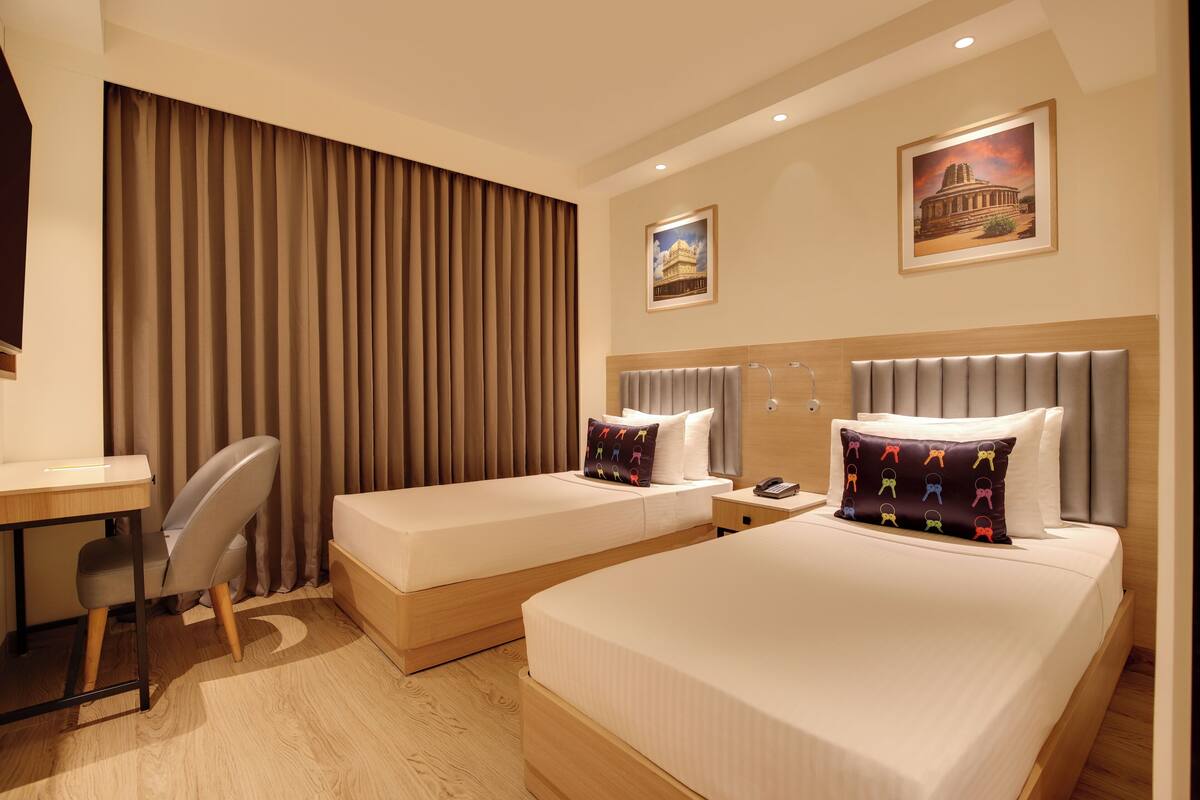 Quarto de hotel com duas camas, uma escrivaninha, uma cadeira e uma cortina.