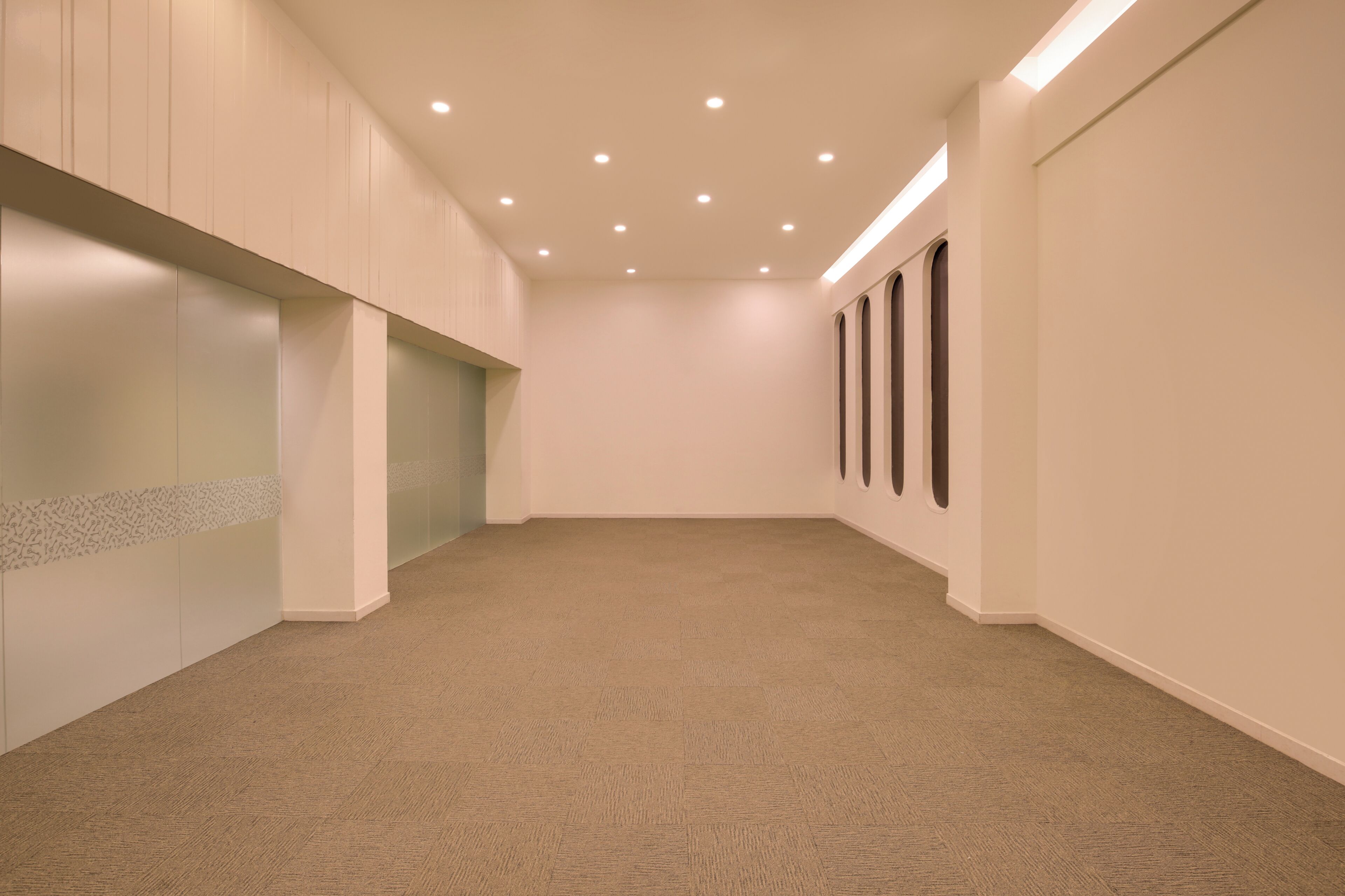 A hallway with carpeted flooring, recessed ceiling lights, and a plain wall with a small window.
