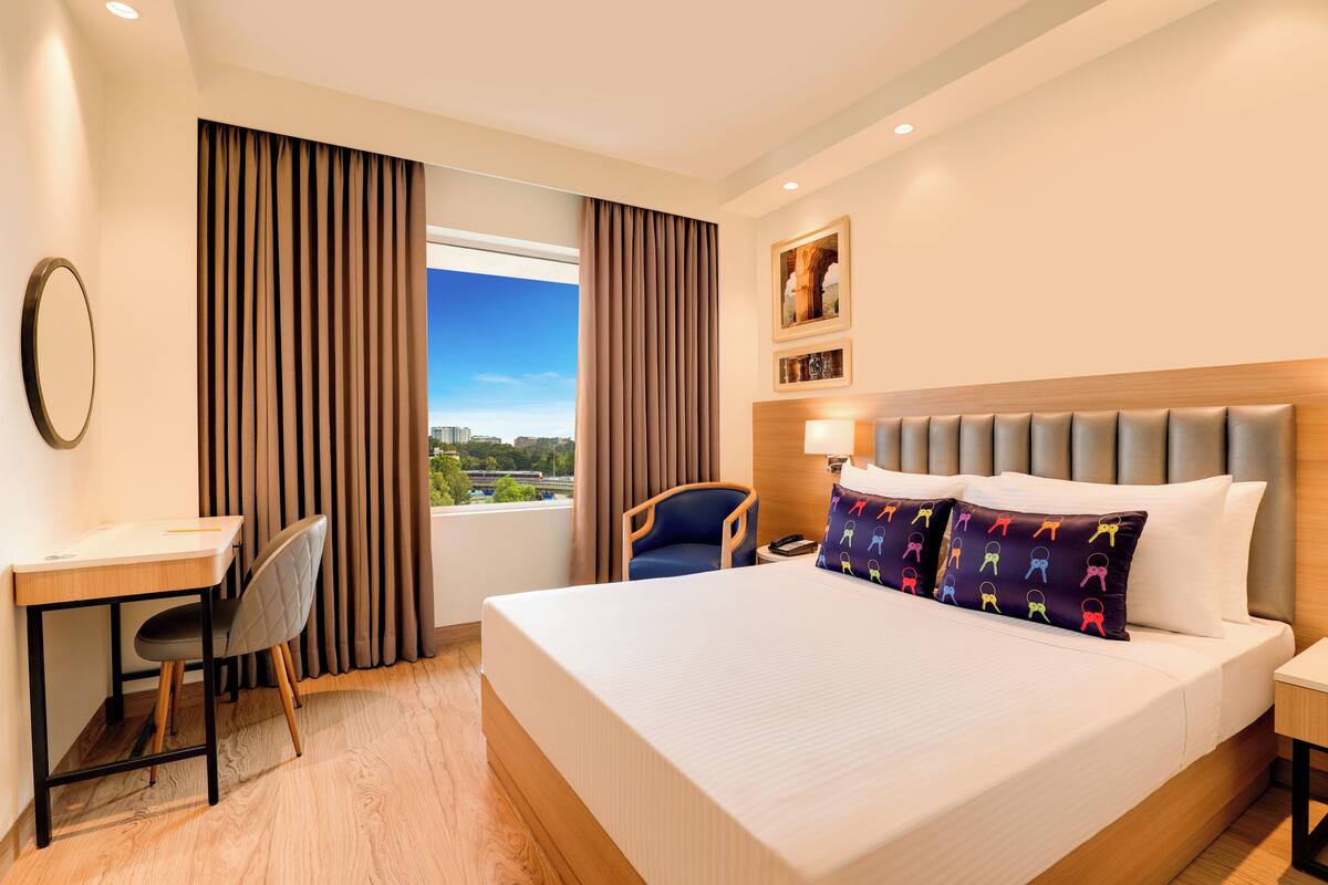 Quarto de hotel com cama, escrivaninha, cadeira e janela com cortinas.