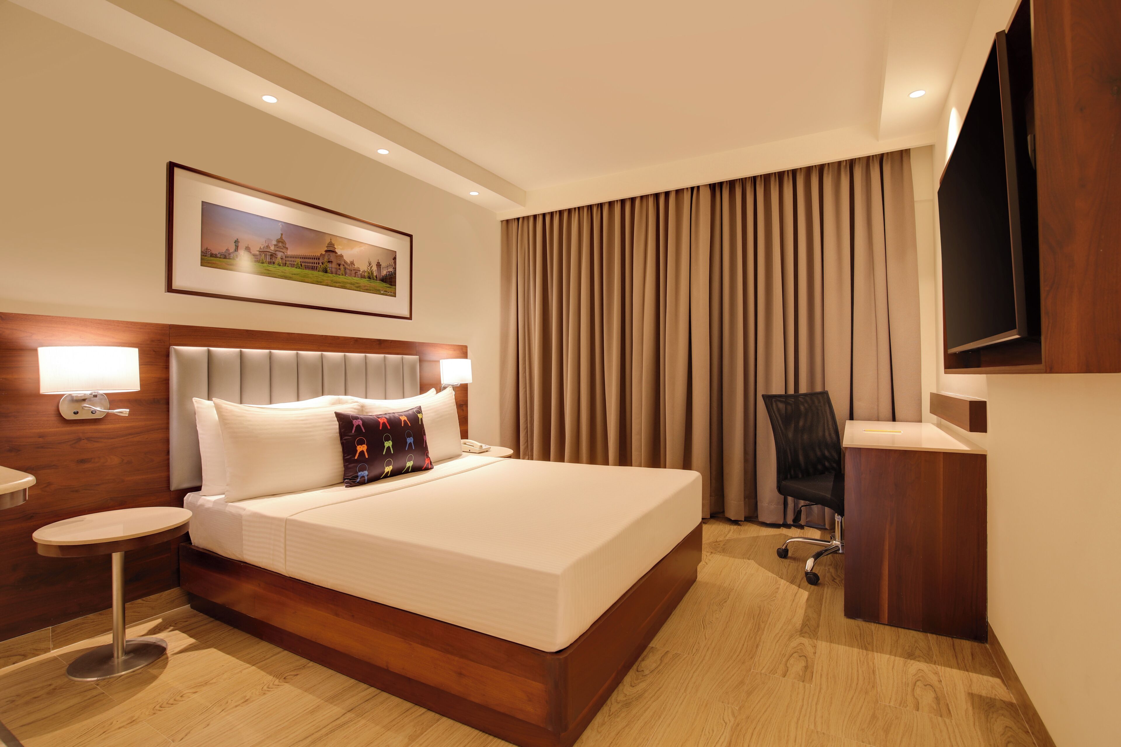 Habitación de hotel con cama, mesita de noche, lámpara, silla, televisor y un cuadro en la pared.