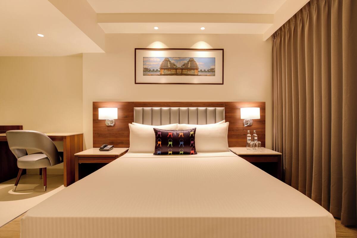 Quarto de hotel com uma cama grande, uma escrivaninha, uma cadeira, uma mesa de cabeceira, um telefone, um travesseiro, um quadro emoldurado na parede e uma cortina.