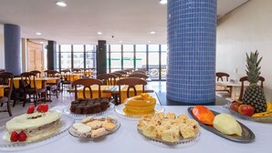A cafeteria with various desserts and fruits on a table, surrounded by chairs and tables. - Confort Hotel (Sao Leopoldo)