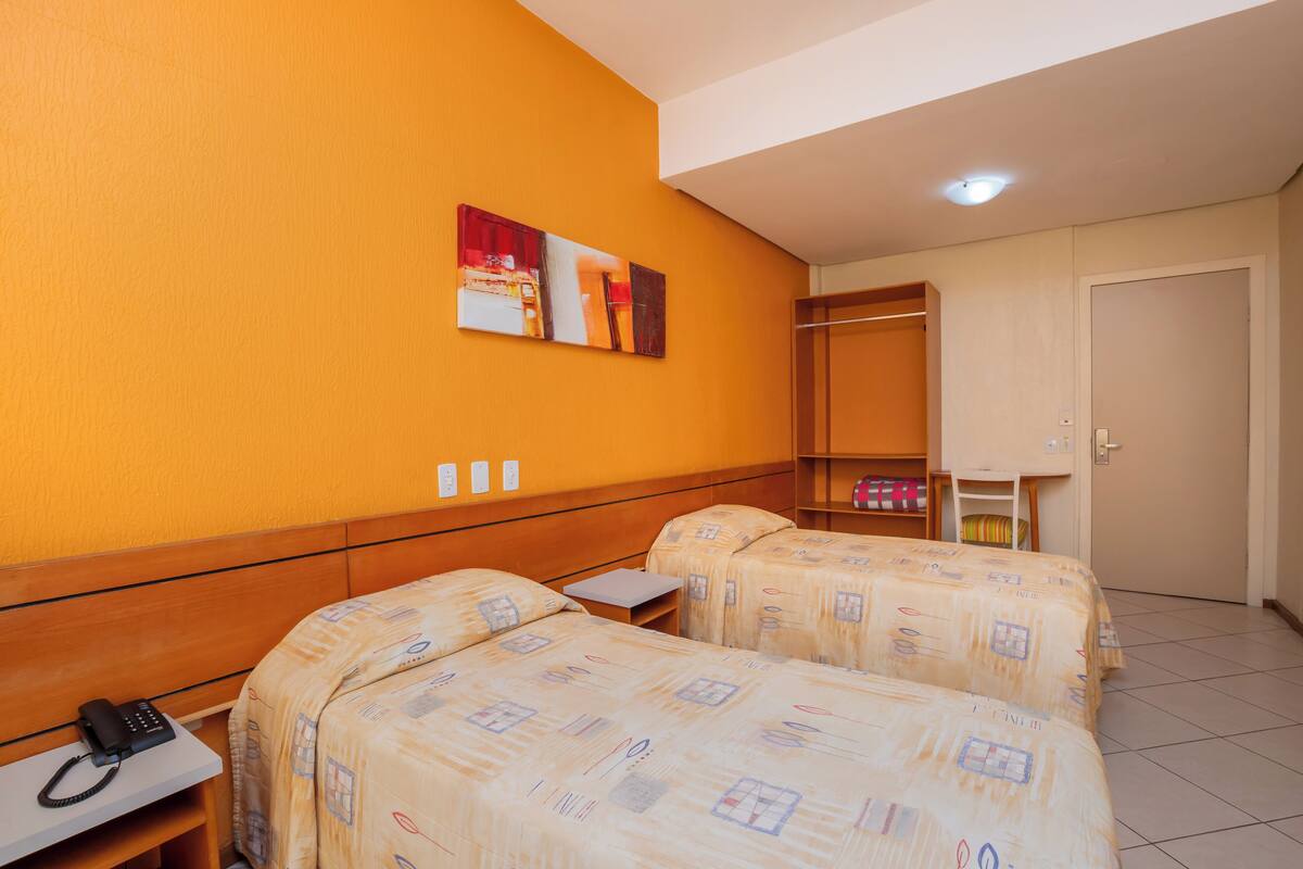 Ein Hotelzimmer mit zwei Betten, einem Schreibtisch mit Telefon, einem Kleiderschrank und einem Gemälde an der Wand.