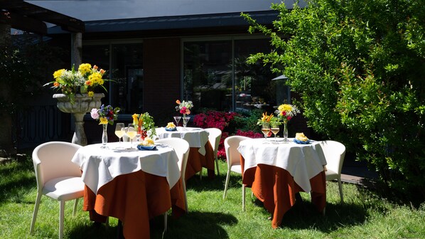 An outdoor dining area with tables set for two, surrounded by lush greenery and a brick building in the background. - Hotel Sunflower (Milan)