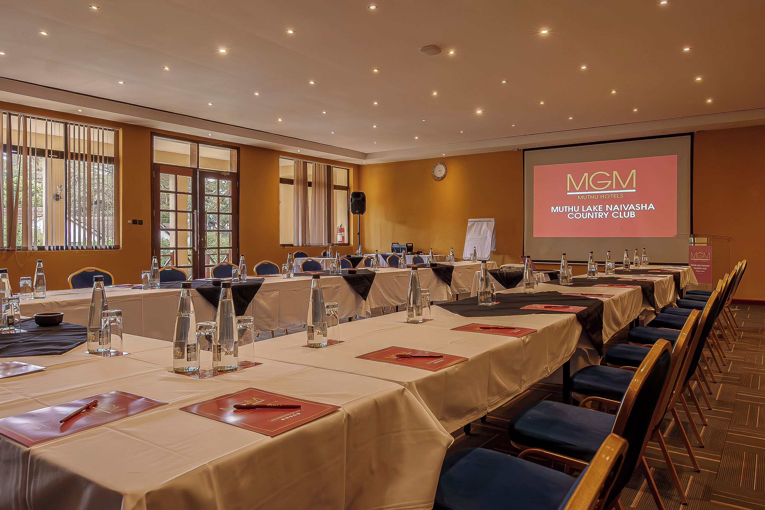 A conference room with long tables, chairs, and a screen displaying the MGM Muthu Lake Naivasha Country Club logo.