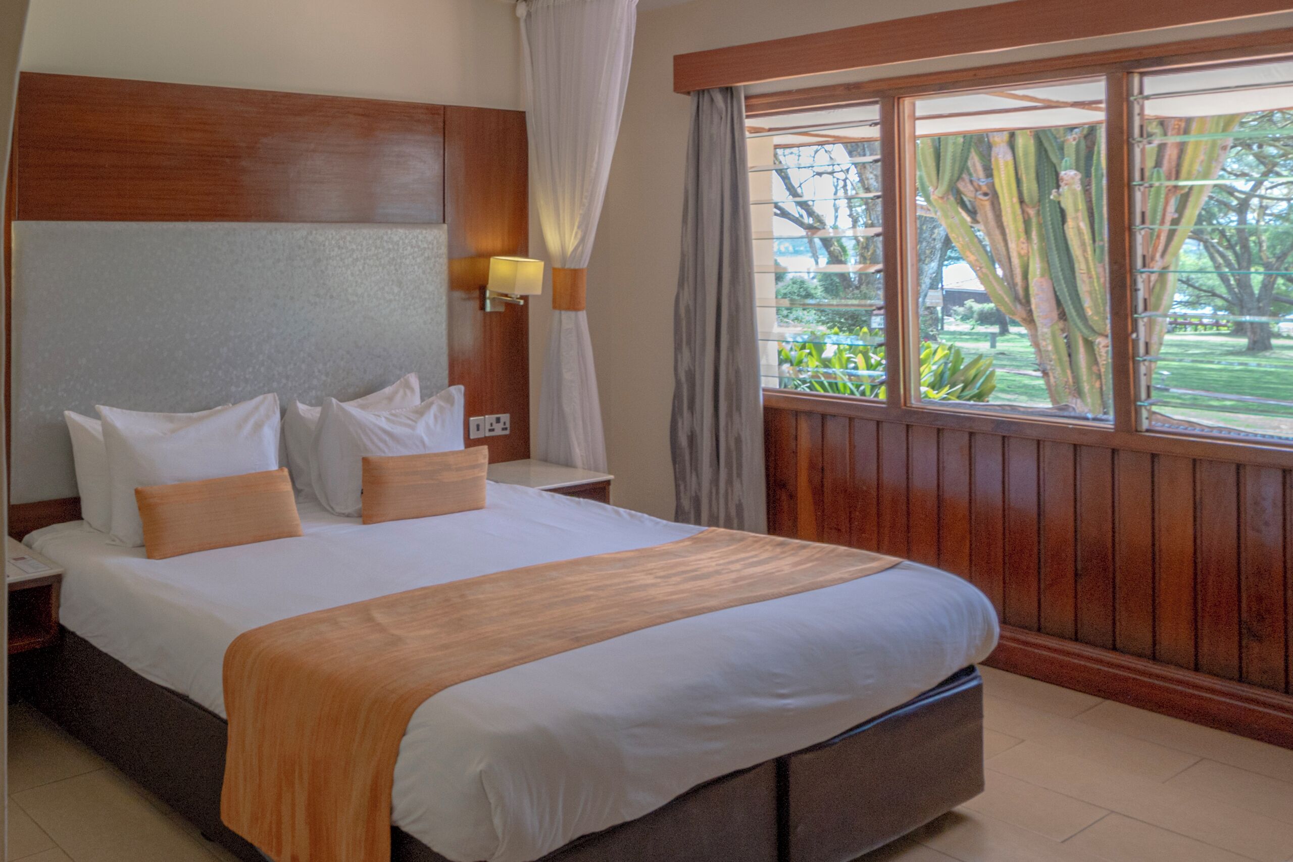 A hotel room with a large bed, wooden headboard, bedside lamps, and a window with a view of greenery.