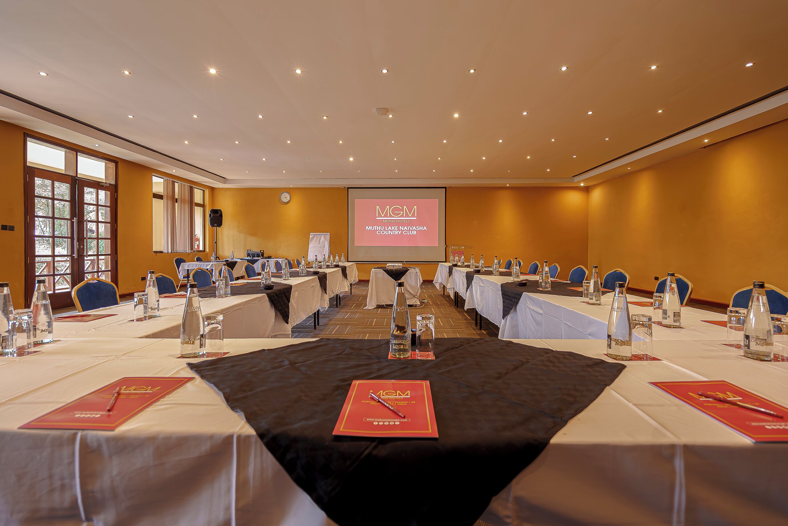 Un salón de conferencias con mesas dispuestas para una reunión, una pantalla mostrando 'MGM MUTHU LAKE NAIVASHA COUNTRY CLUB', y un ventanal grande a la izquierda.