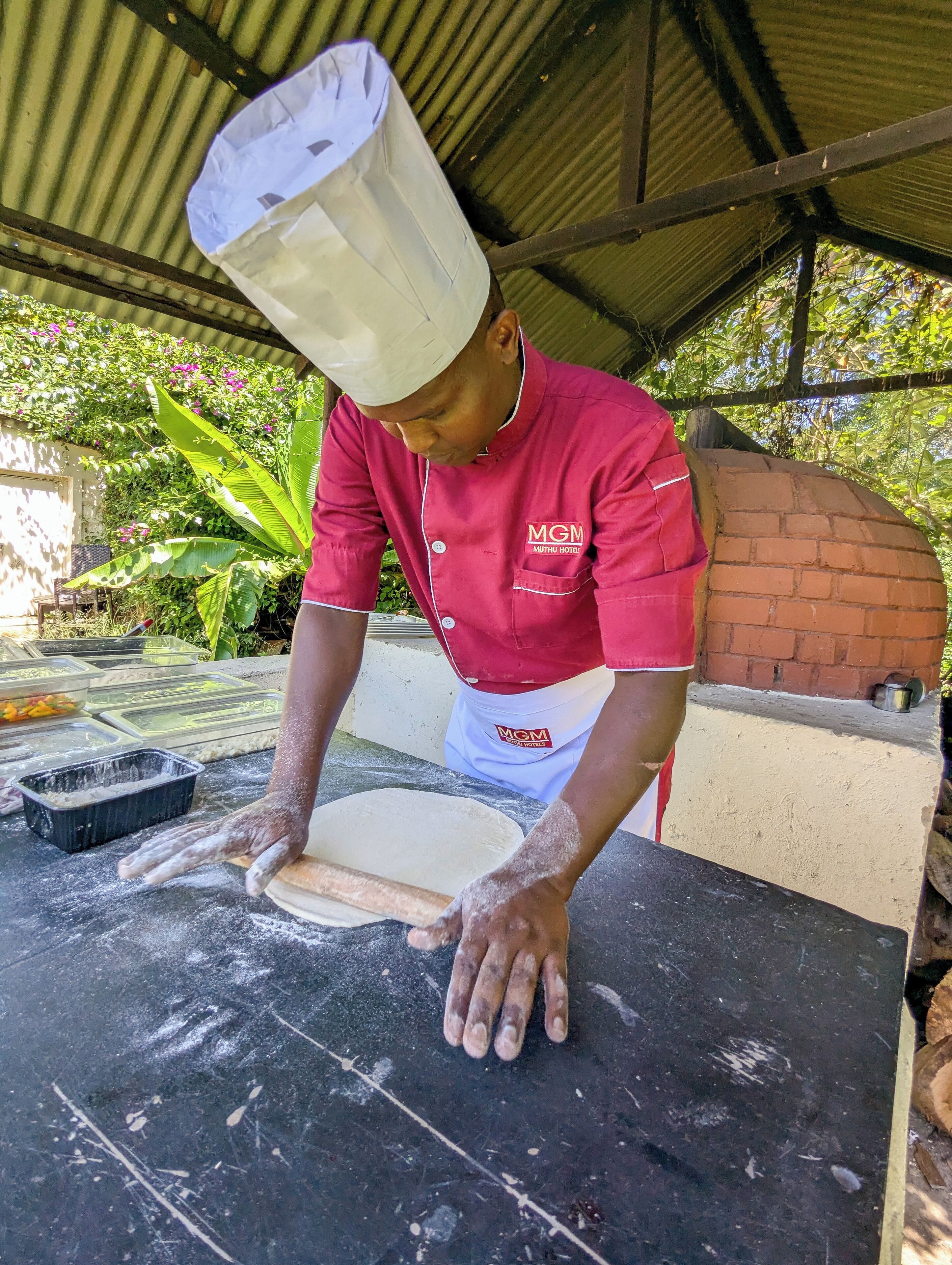 A chef preparing dough on a marble countertop under a corrugated metal roof, with a brick oven and greenery in the background.