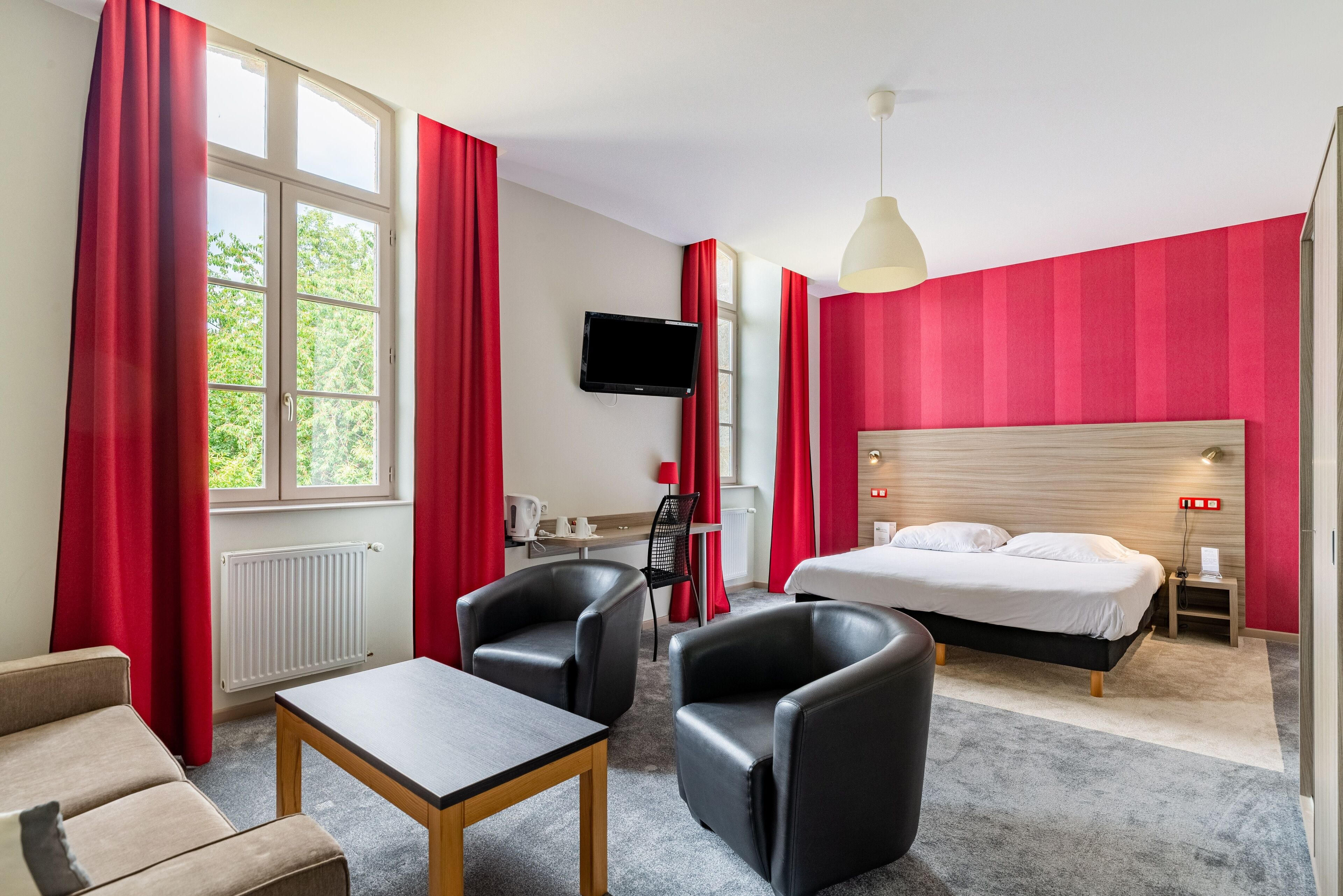 Habitación de hotel con cama, escritorio, silla, televisor y ventana con cortinas rojas.