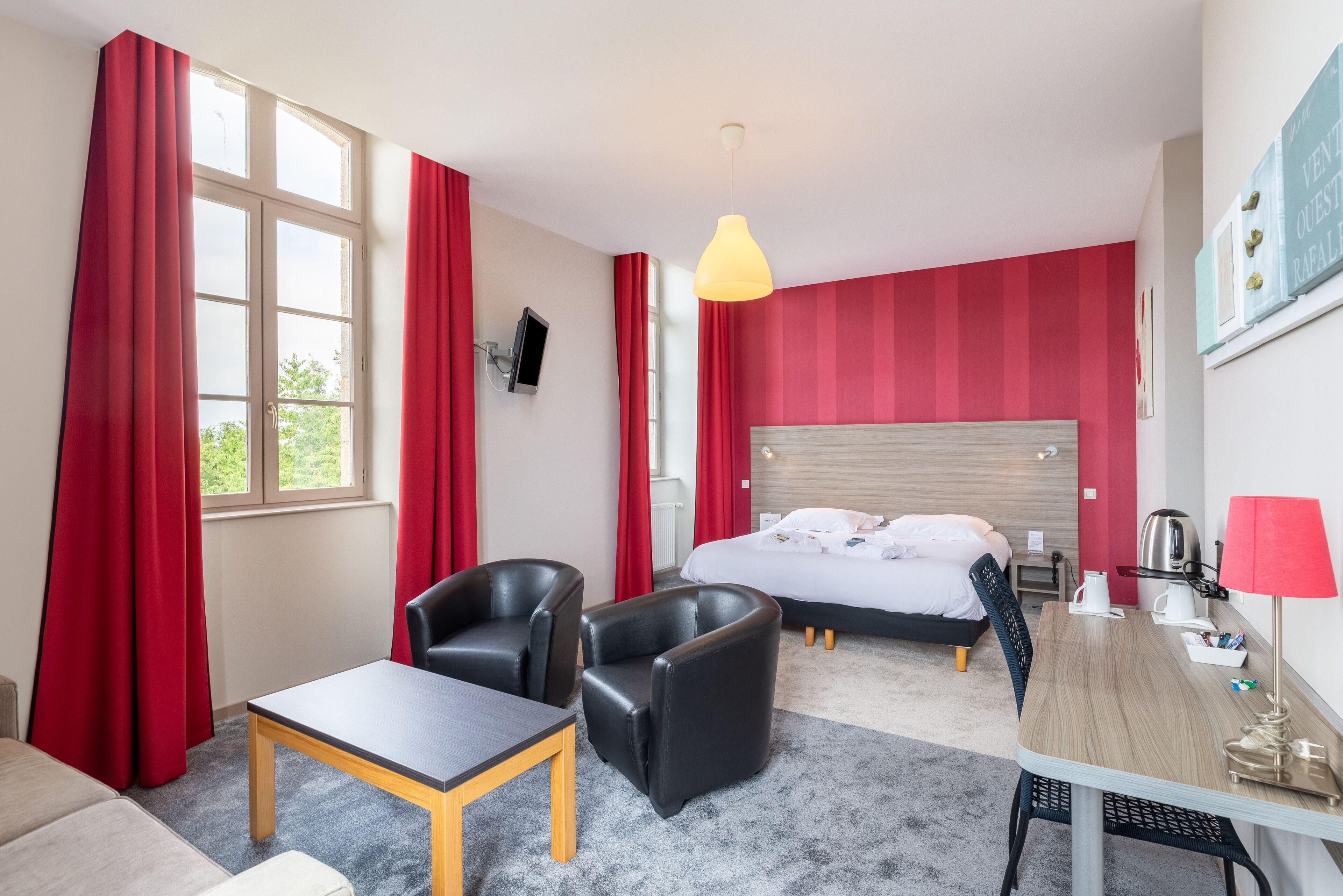 A hotel room with a bed, two chairs, a table, a desk, a lamp, and a TV.
