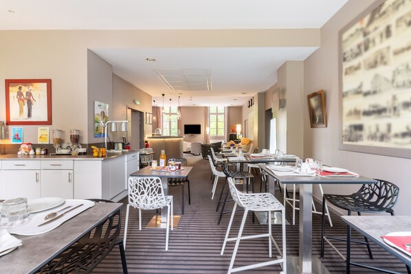 Un comedor moderno de hotel con una larga mesa común, mesas individuales y una zona de cocina al fondo.