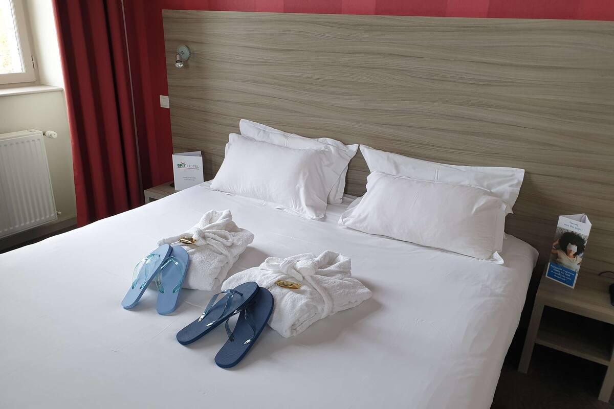 A hotel room with a neatly made bed, white linens, and a wooden headboard.