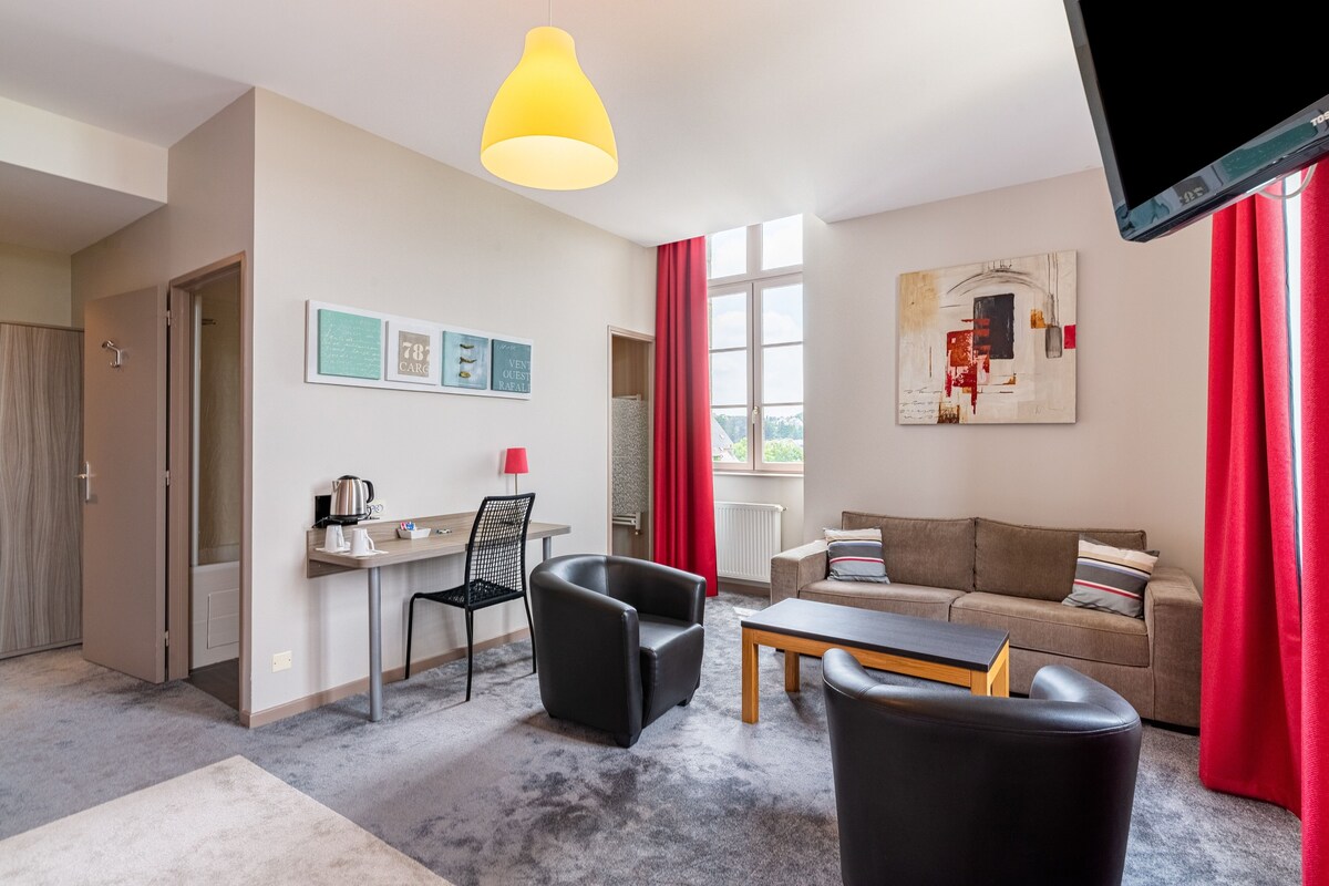 Una sala de estar moderna con un sofá, una mesa de centro y un televisor. Hay un comedor con una silla y una mesa, y una cocina con una tetera y una cafetera.
