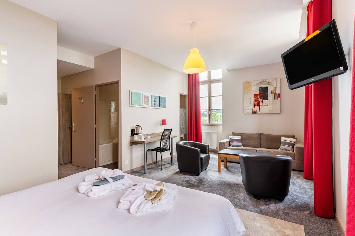 A hotel room with a bed, a desk, a chair, a sofa, a TV, and a window.