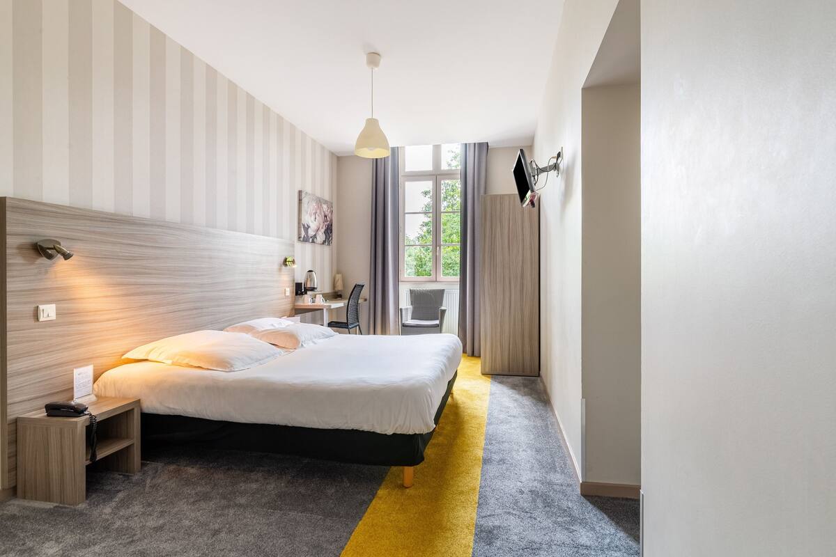 A modern hotel room with a large bed, a desk, a chair, and a view of the outdoors.