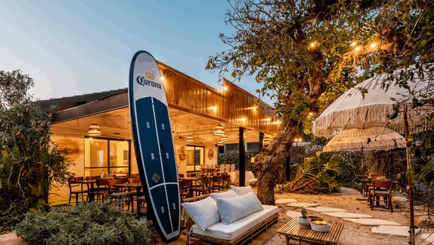 An outdoor dining area with wooden furniture, a surfboard, and a thatched umbrella. - Dolphin Village - Sea Resort (Mateh Asher)