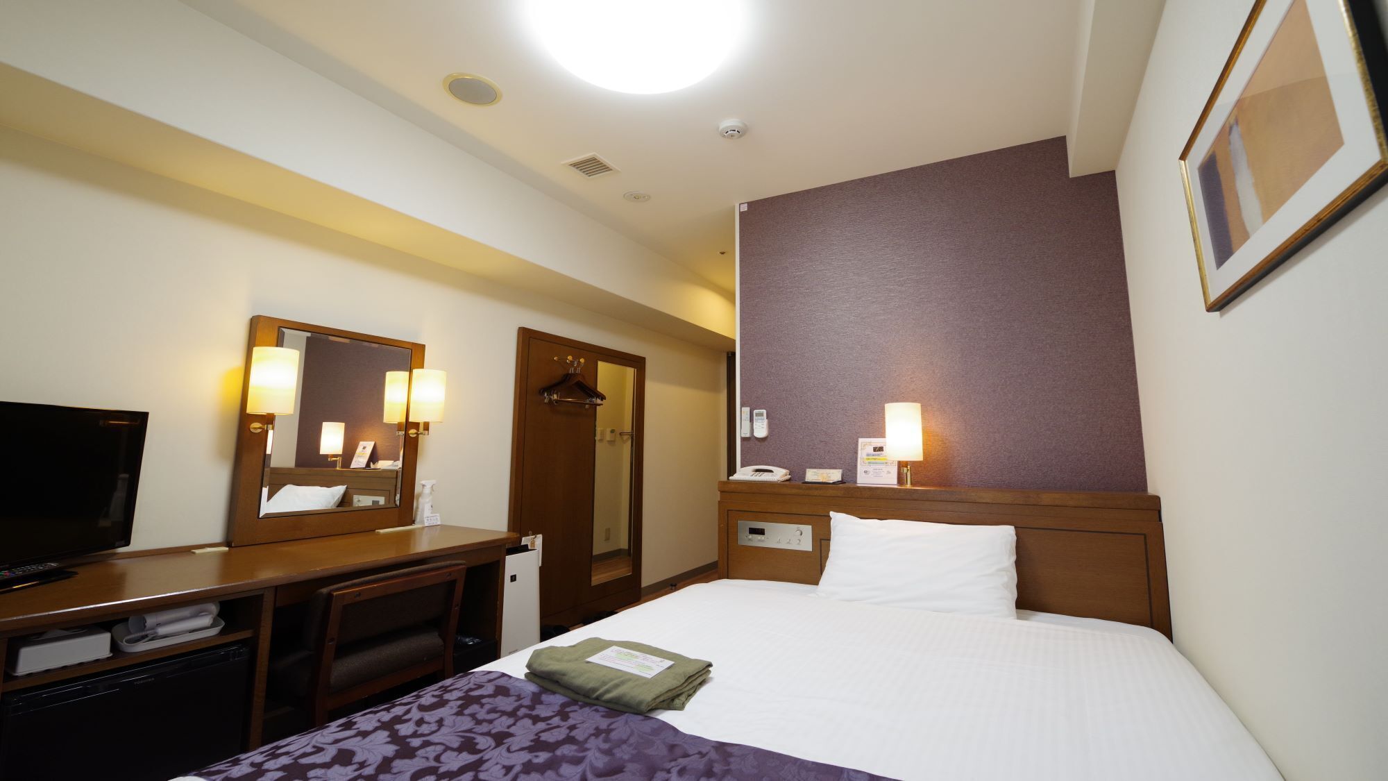 A hotel room with a bed, bedside table, TV, and a mirror.