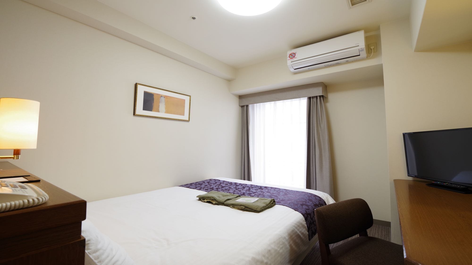 A hotel room with a bed, a desk with a lamp, a television, and an air conditioning unit.