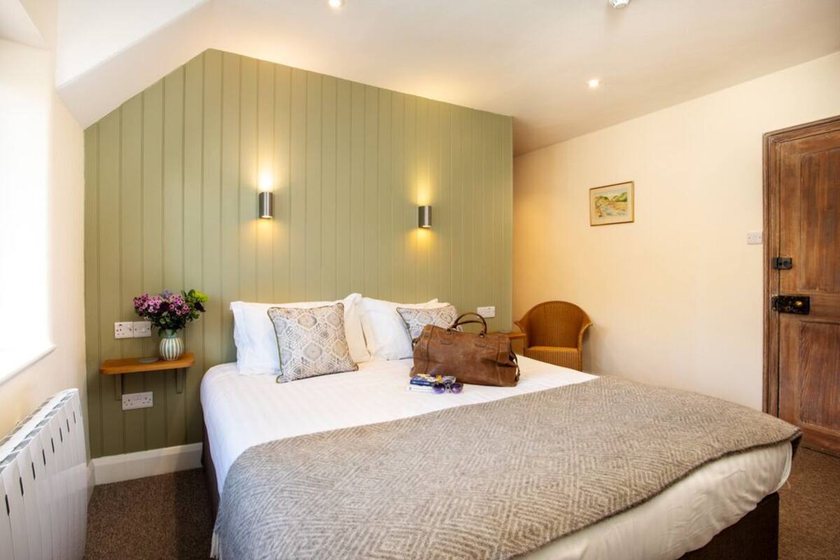 A bedroom with a bed, pillows, a bedside table with a vase of flowers, a chair, and a wooden door.