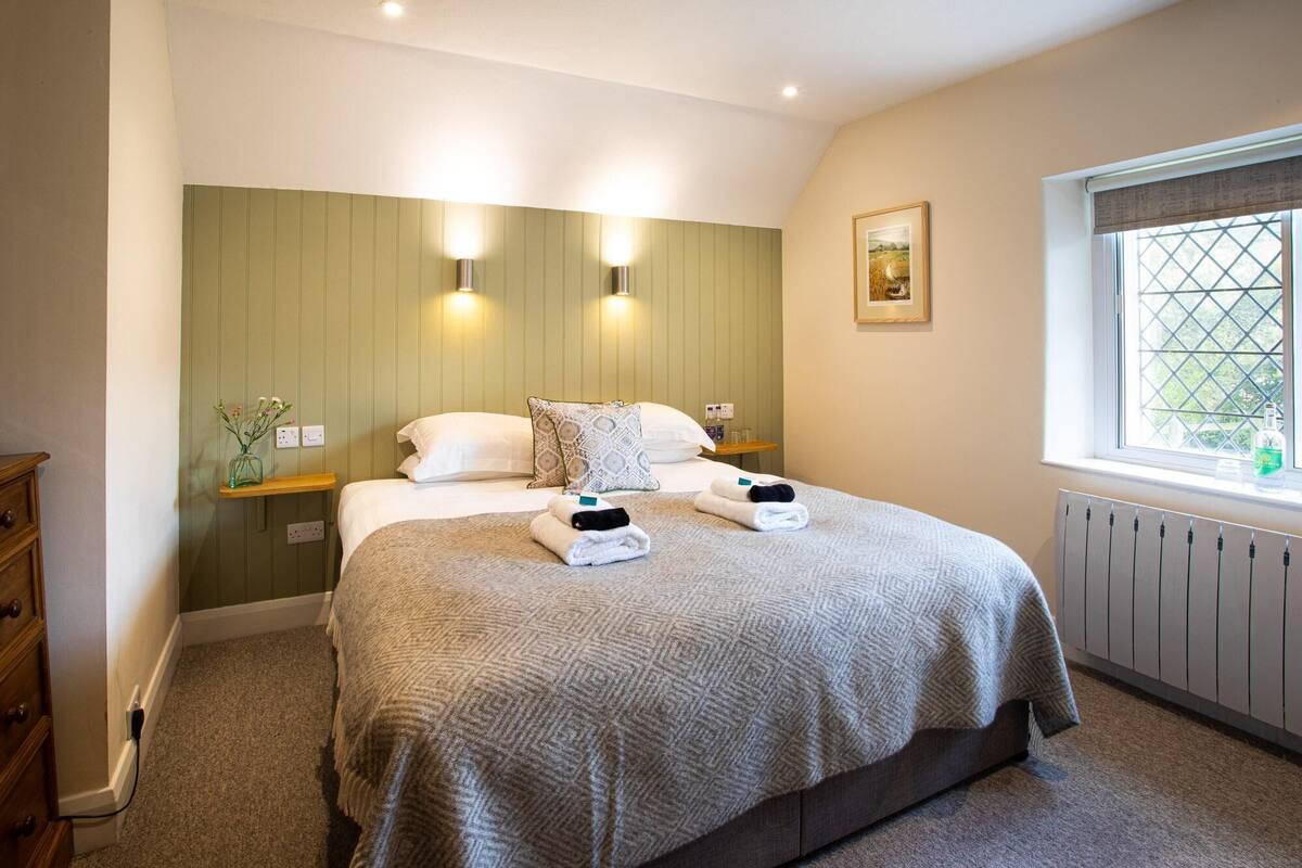 A neatly made bed with a patterned blanket, two bedside lamps, a small bedside table, and a window with a view of greenery.