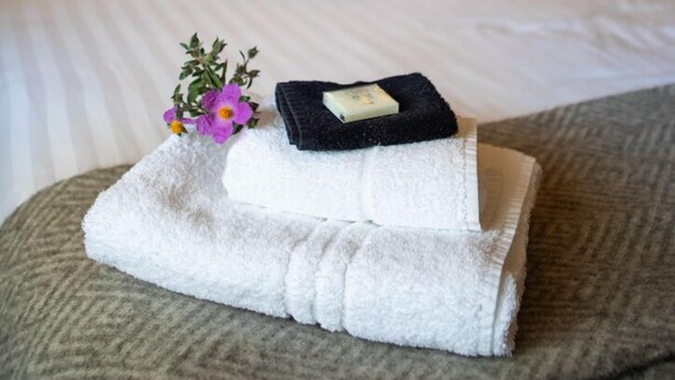 Una cama bien tendida con sábanas blancas, una toalla doblada, un albornoz negro y un pequeño florero con flores sobre un reposapiés con textura.