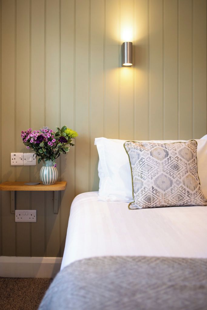 A hotel room with a bed, a bedside table with a vase of flowers, and a wall-mounted light fixture.