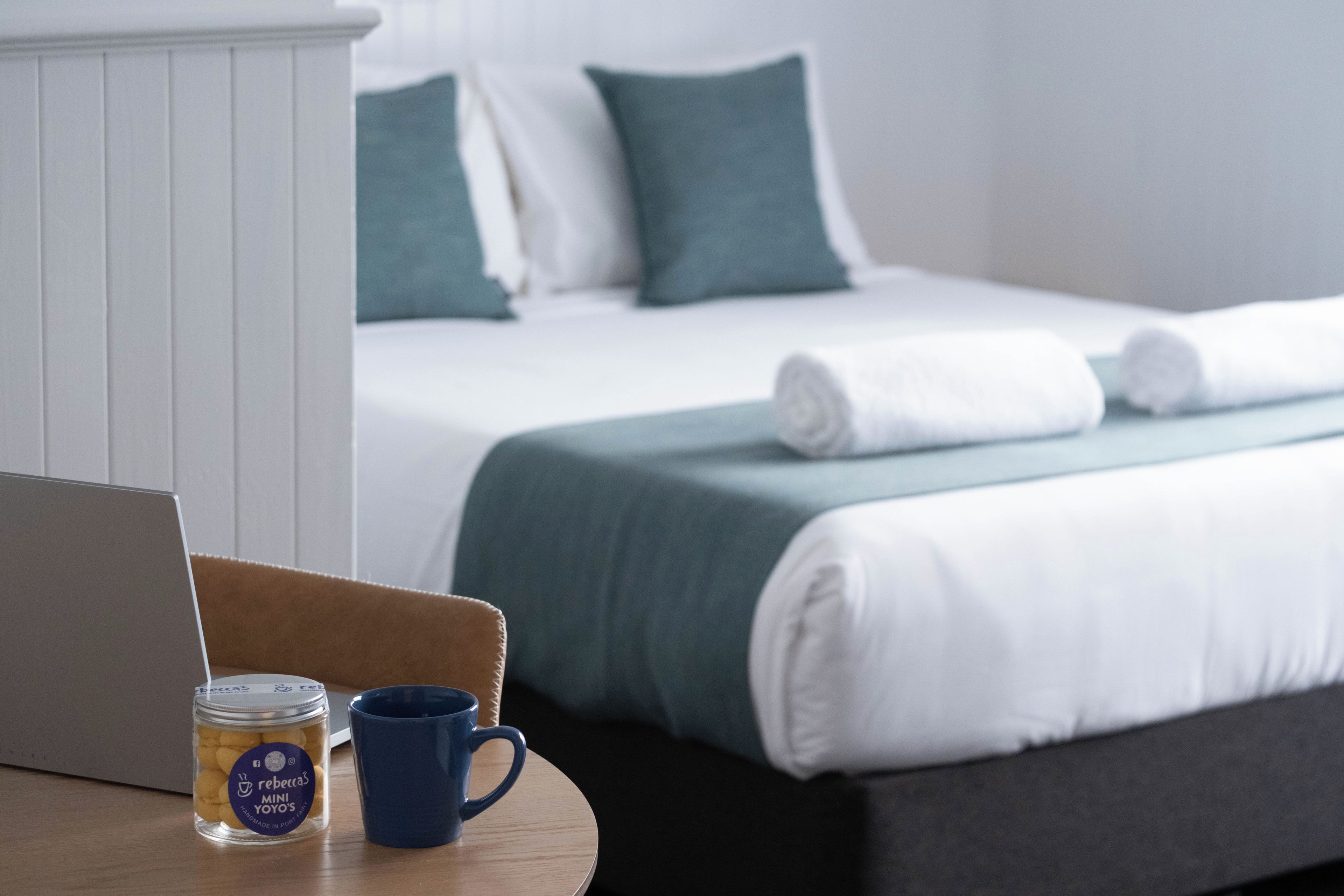 A neatly made bed with white and green bedding, a laptop, a mug, and a jar of cookies on a wooden table.