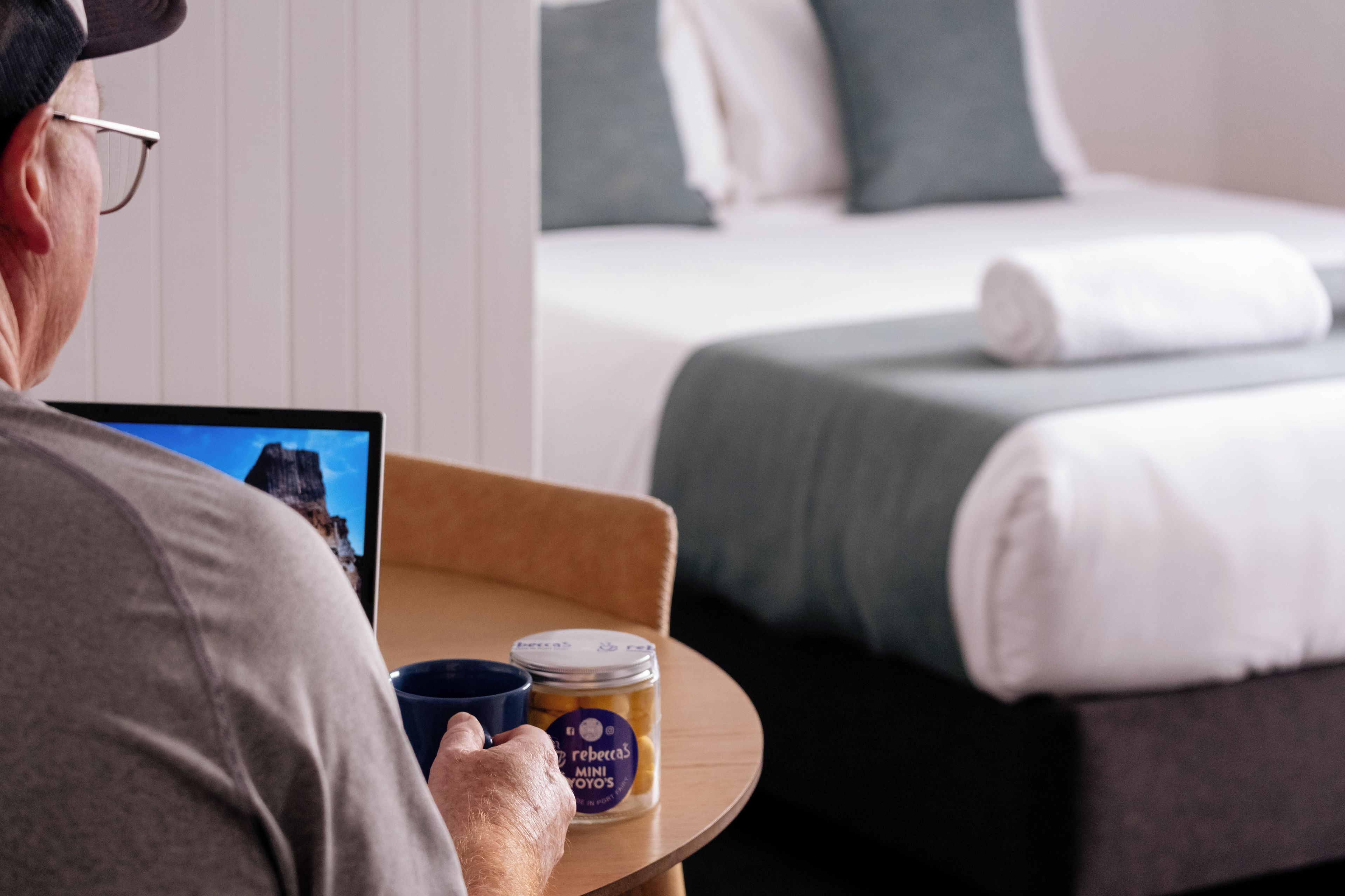 A person seated at a table with a tablet, a blue mug, and a jar of cookies. In the background, there is a bed with a green pillow and a white blanket.