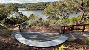 Eine Holztreppe führt zu einer Aussichtsplattform mit Metallgeländer, umgeben von Bäumen und mit Blick auf ein Gewässer.
