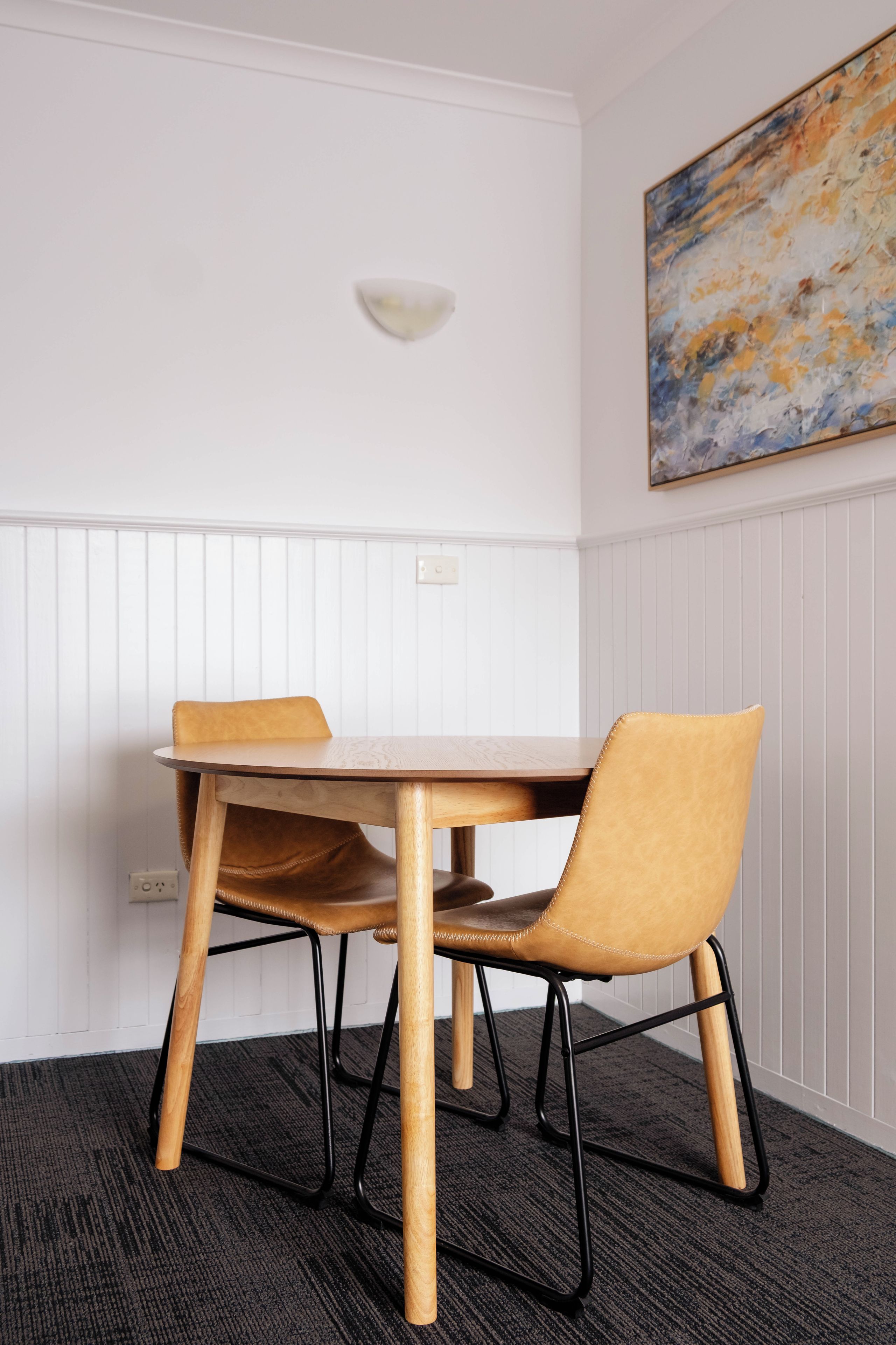 A dining area with a wooden table and two chairs, a light fixture on the wall, and a painting above the table.