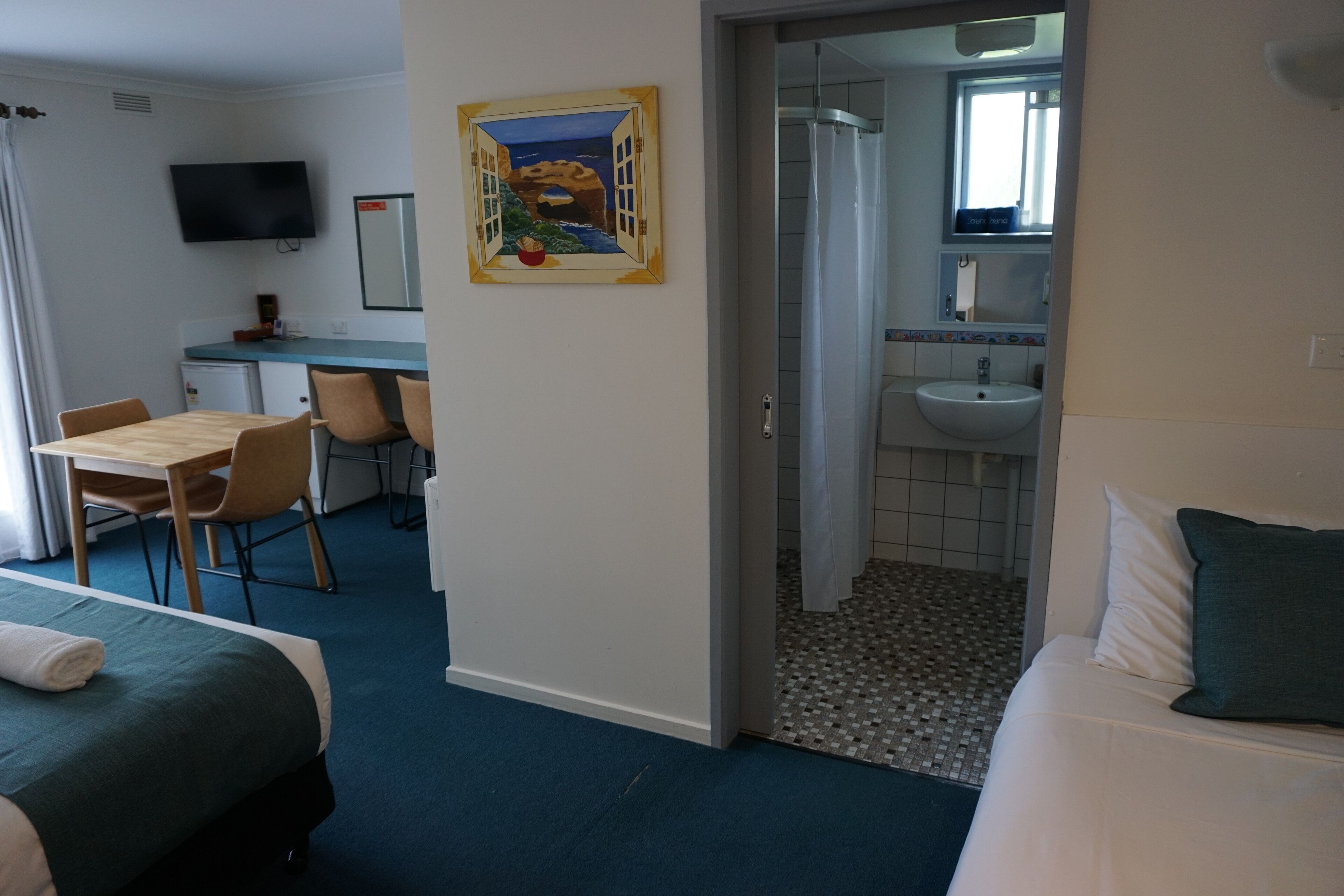 A hotel room with a bed, a desk, and a chair. There is a painting on the wall and a bathroom visible through an open door.