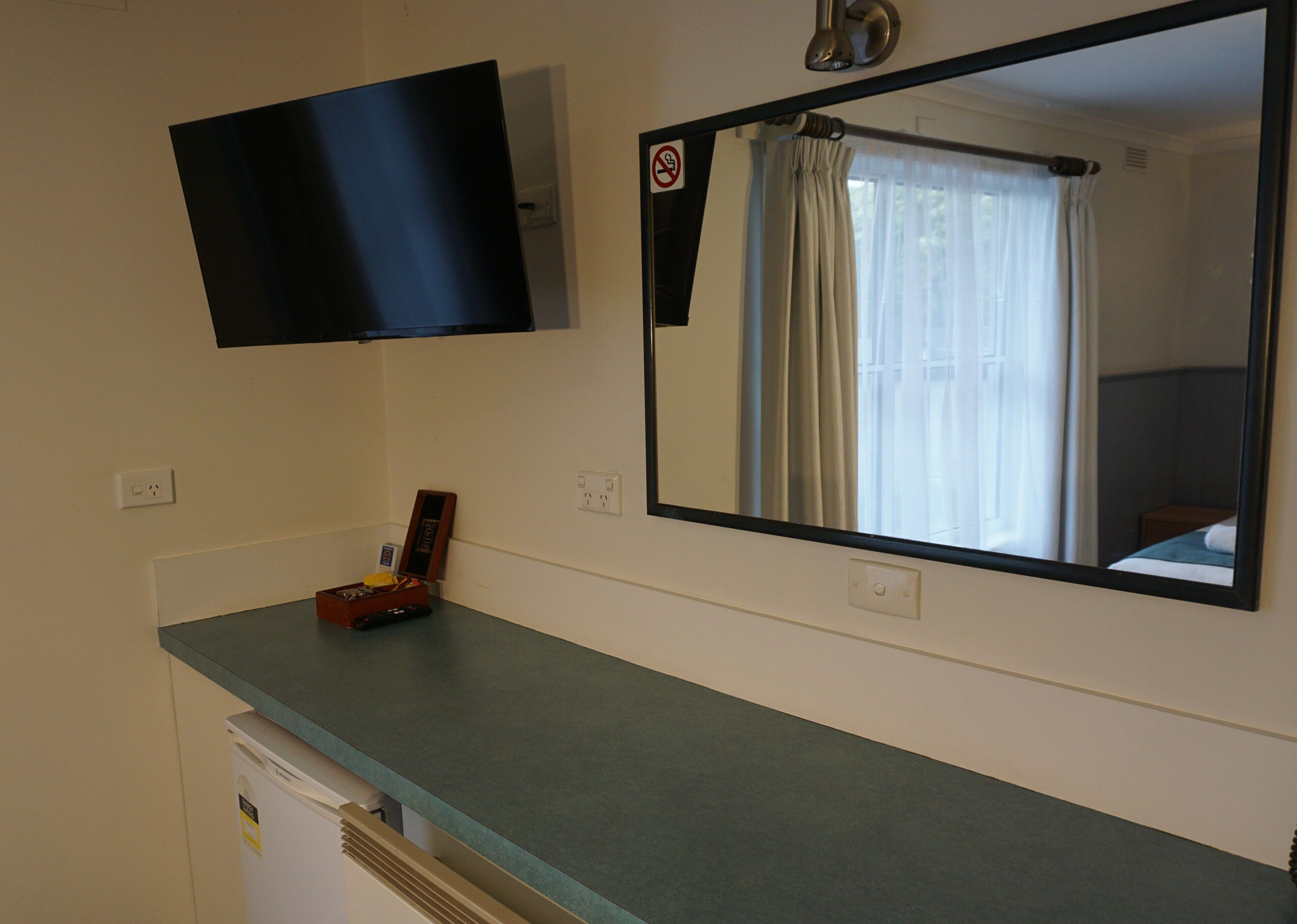 Una habitación con un televisor de pantalla plana montado en la pared, un espejo que refleja una ventana con cortinas, una caja de madera pequeña sobre el mostrador y un electrodoméstico blanco debajo.