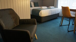 Ein Hotelzimmer mit einem Bett, einem dunklen Sessel, einem Holzhocker und einem blauen Teppich.