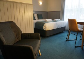 Una habitación de hotel con una cama, un sillón oscuro, una silla de madera y una alfombra azul.