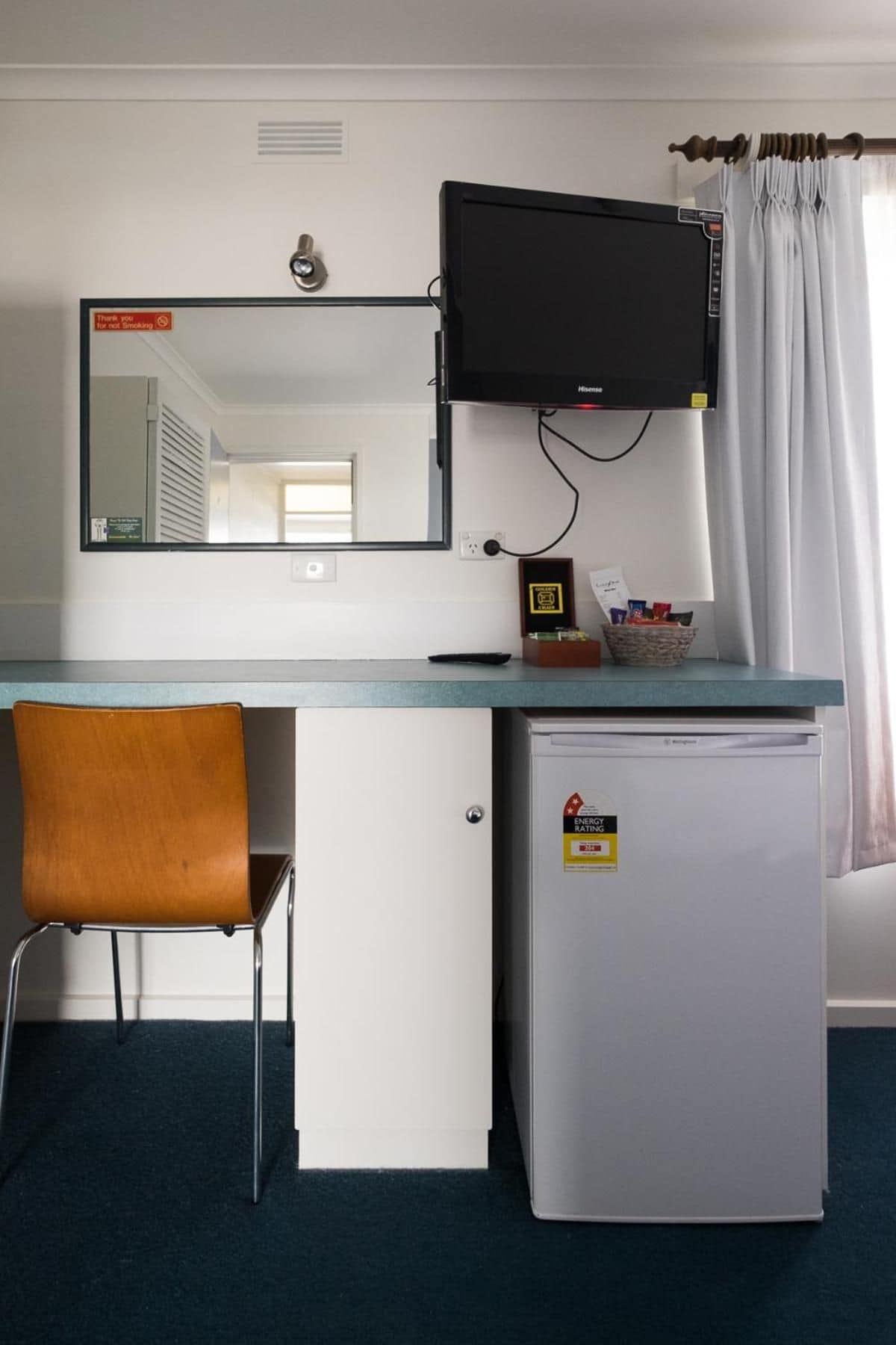 Een kleine hotelkamer met een bureau, een stoel, een televisie boven het bureau, een minibar en een raam met gordijnen.