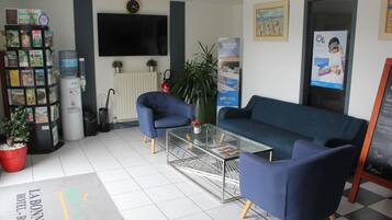 Zona de recepció d'hotel amb taulell de recepció, un sofà blau, taula de cafè de vidre, una planta en test, un dispensador d'aigua i una gran pantalla de televisió.