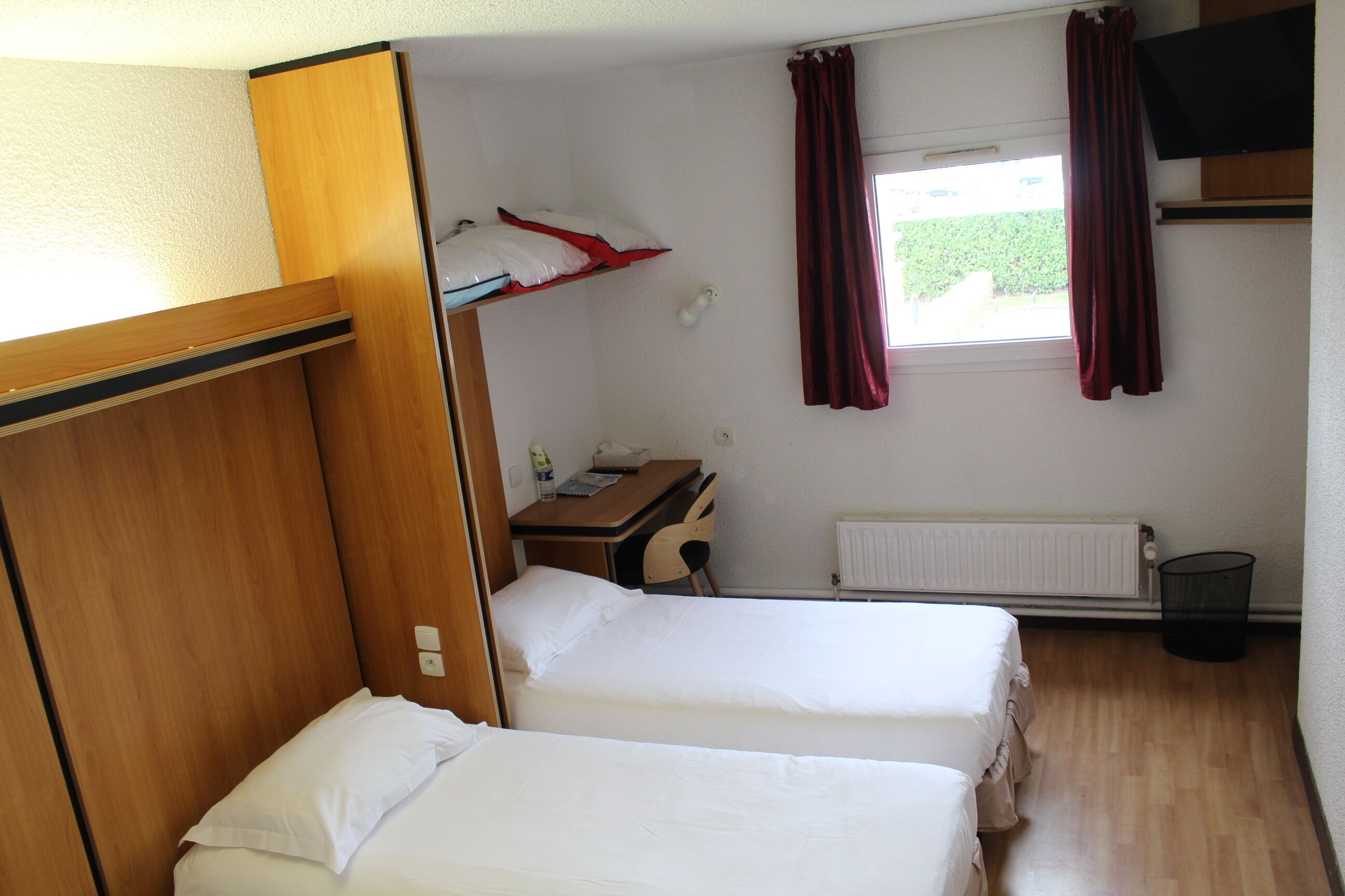 A compact room with two beds, a desk, a chair, a radiator, and a window with curtains.