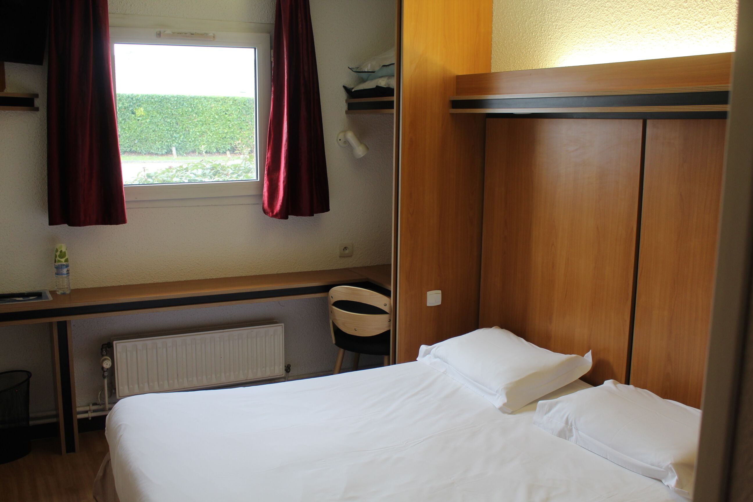 A hotel room with a bed, a window with curtains, a radiator, a chair, and a wooden wardrobe.