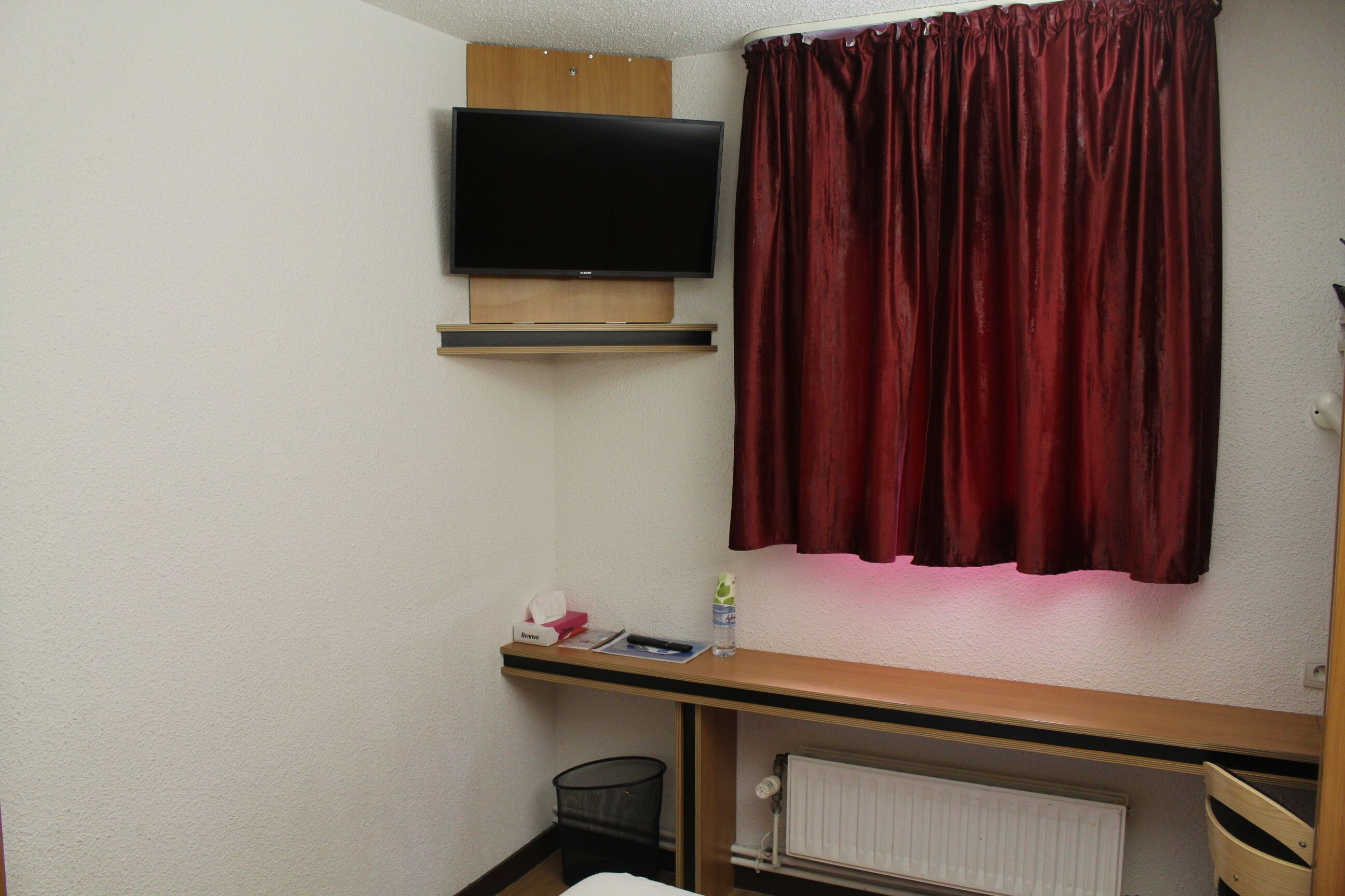 A small room with a television mounted on the wall, a red curtain, a wooden shelf with a tissue box and a bottle, a radiator, and a black trash bin.