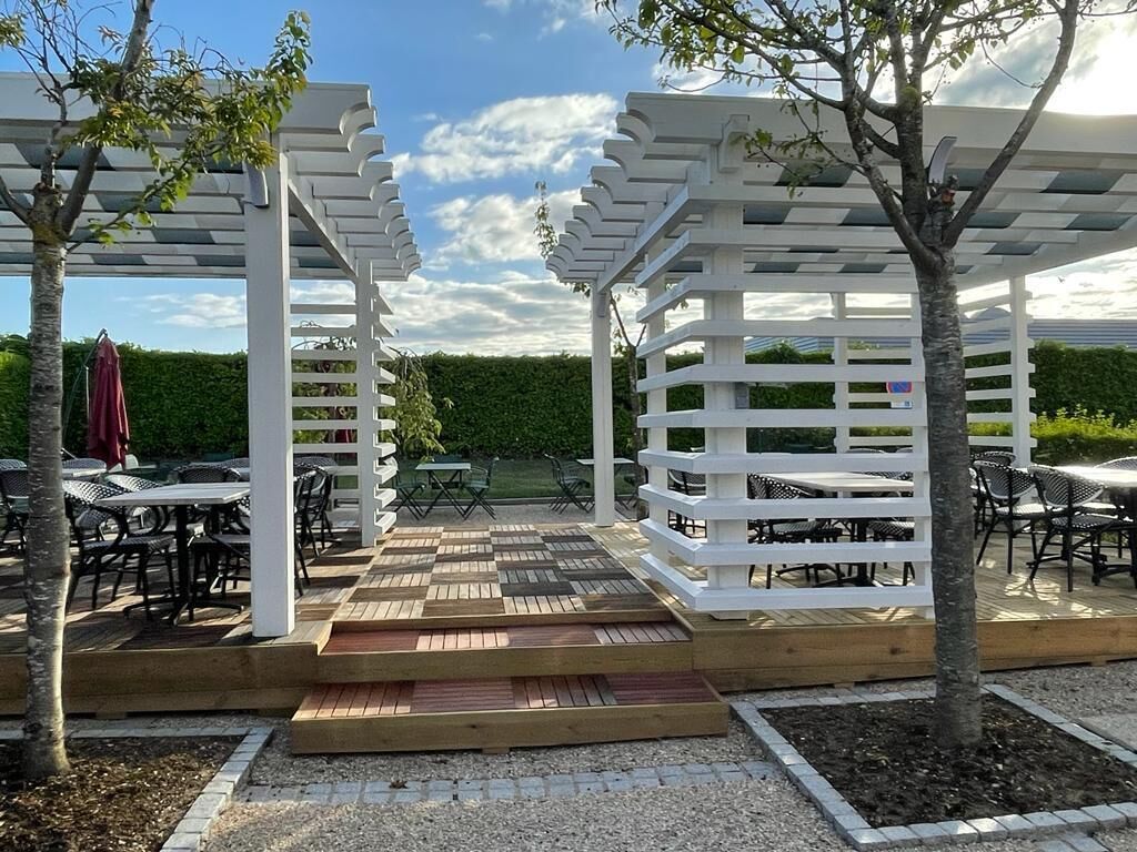 Ett takförsett utomhusområde med vita pergola, trägolv samt bord och stolar.