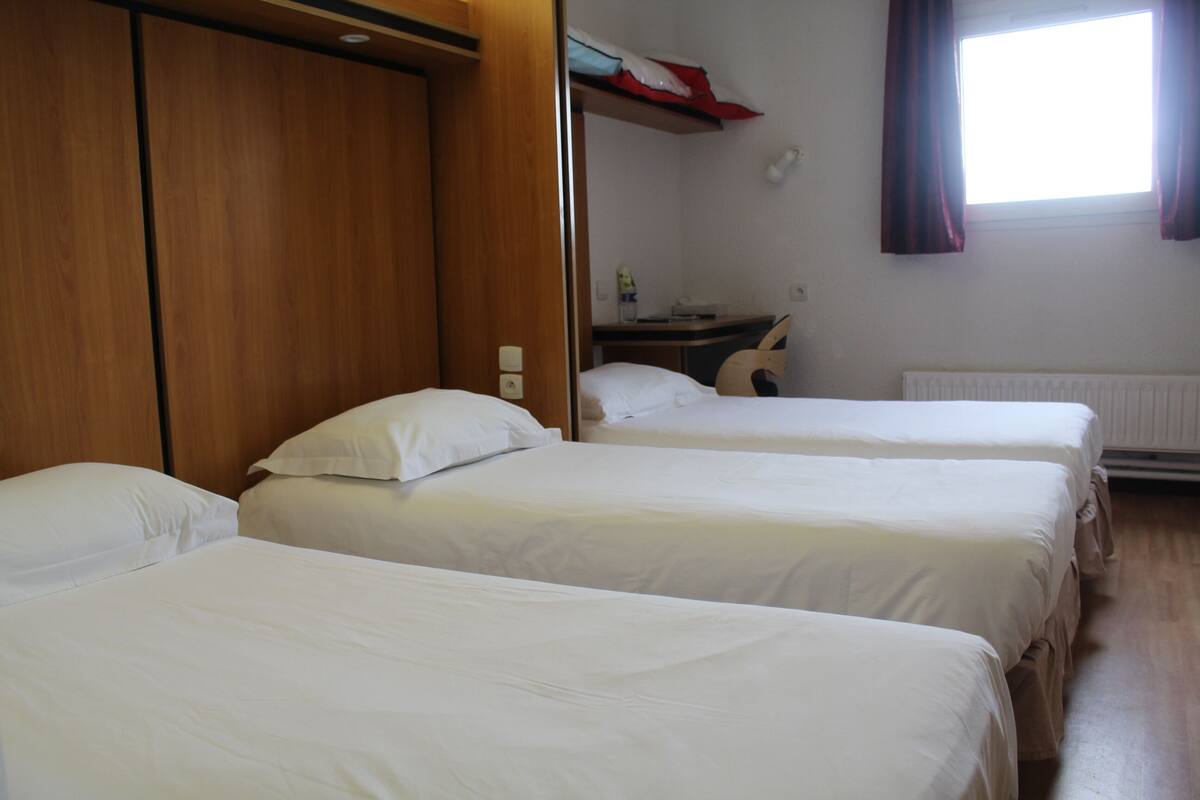 Habitació d'hotel amb tres llits individuals, un armari de fusta, escriptori amb cadira i finestra amb cortines.