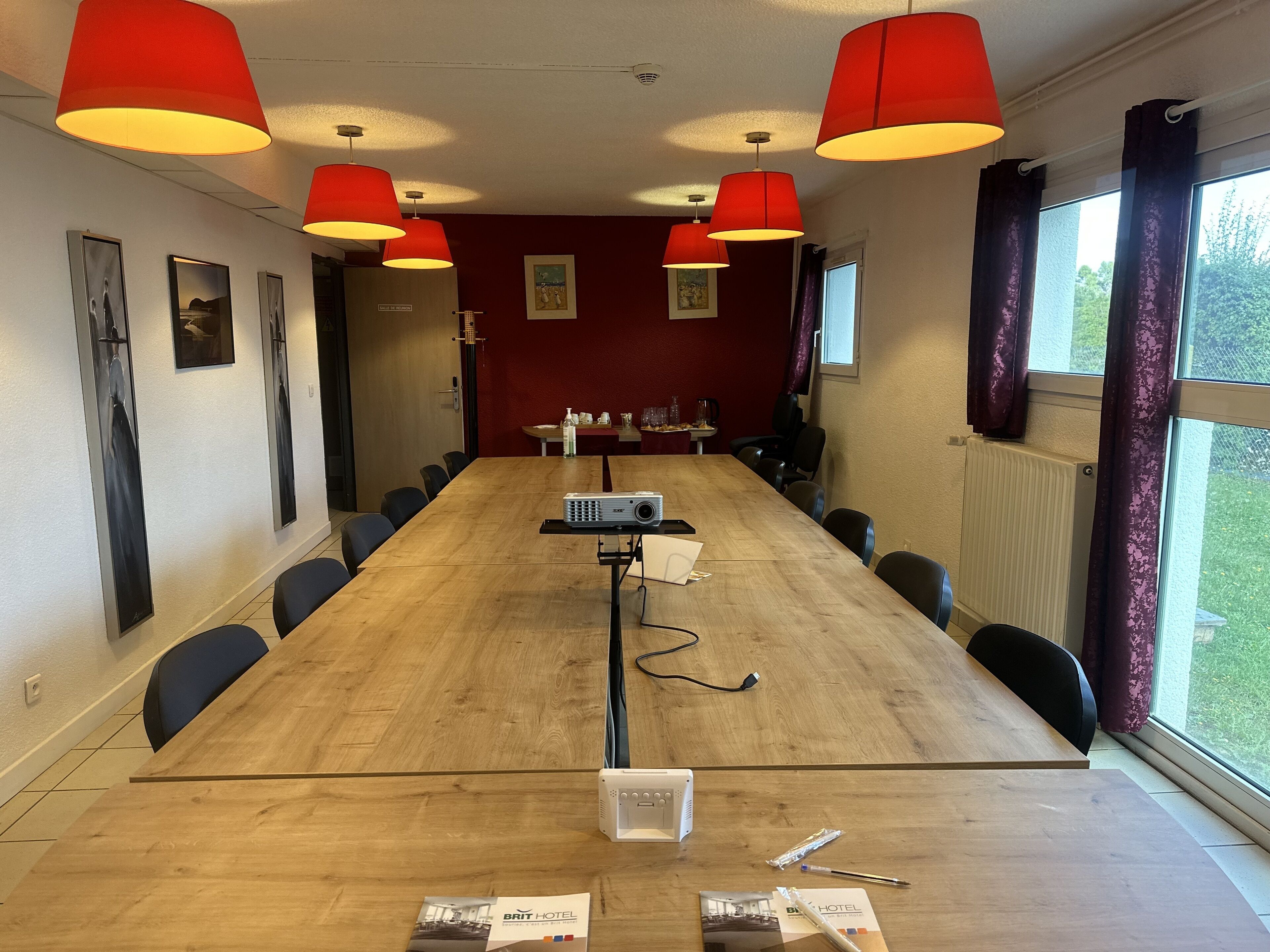 Une salle de conférence avec une longue table en bois, des chaises noires, des lampes suspendues rouges et un projecteur.