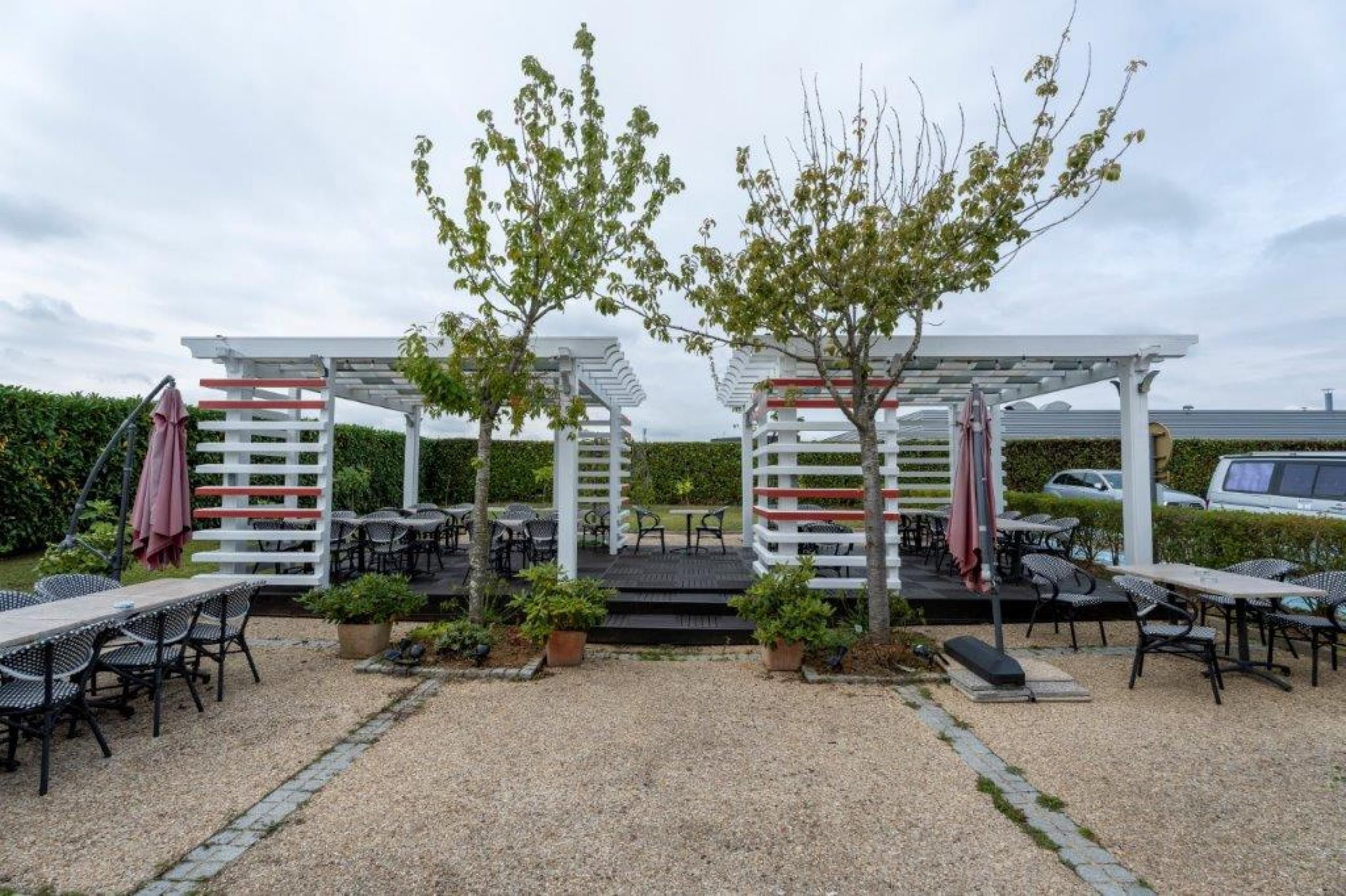 Un espace extérieur aménagé en terrasse, avec des tables et des chaises, recouvert de pergolas blanches agrémentées de touches de rouge, et entouré de verdure.
