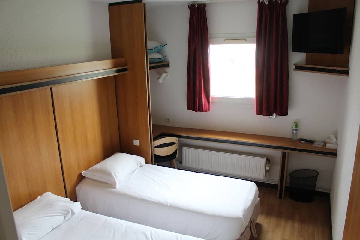 Habitació d'hotel senzilla i compacta amb dos llits individuals, un capçal de llit de fusta, un escriptori petit amb una cadira, un televisor fixat a la paret i una finestra amb cortines vermelles.
