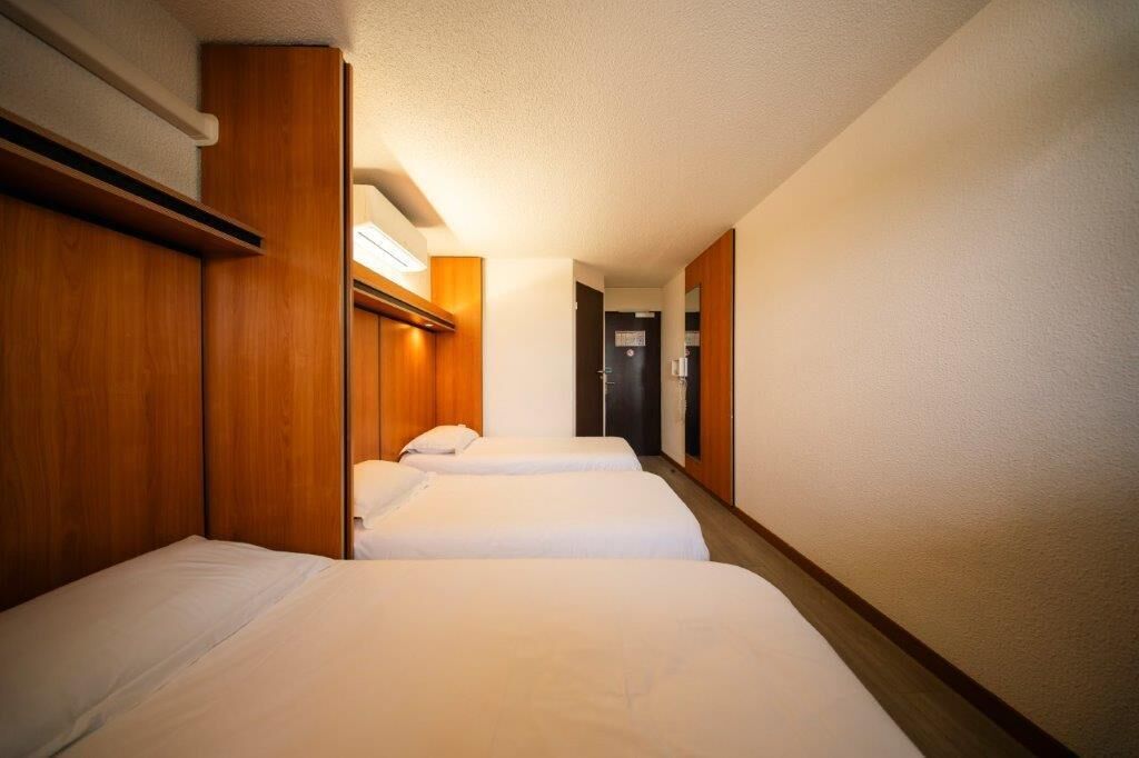 A hotel room with two beds, wooden headboards, and a hallway leading to a door.