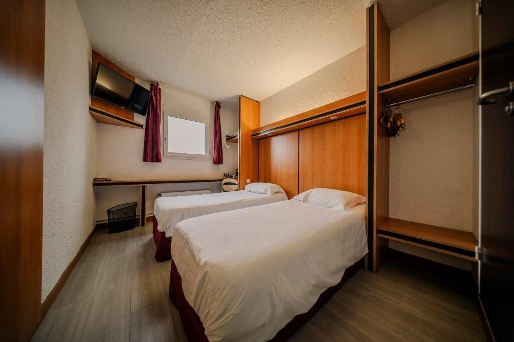 Ett hotellrum med två sängar, en tv som är monterad på väggen, ett fönster med gardiner och en garderob i trä.