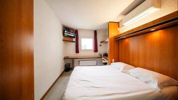 A compact hotel room with a single bed, a small desk, a television, and a window with curtains.